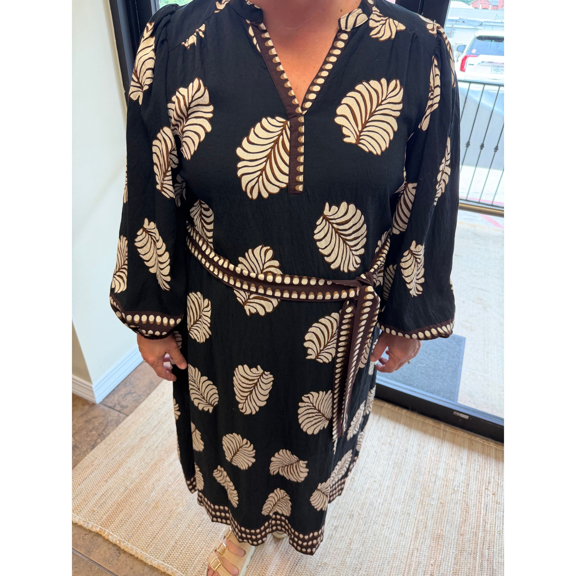 A woman wearing a black wrap-style dress with beige leaf patterns and brown trim, standing indoors near a window.