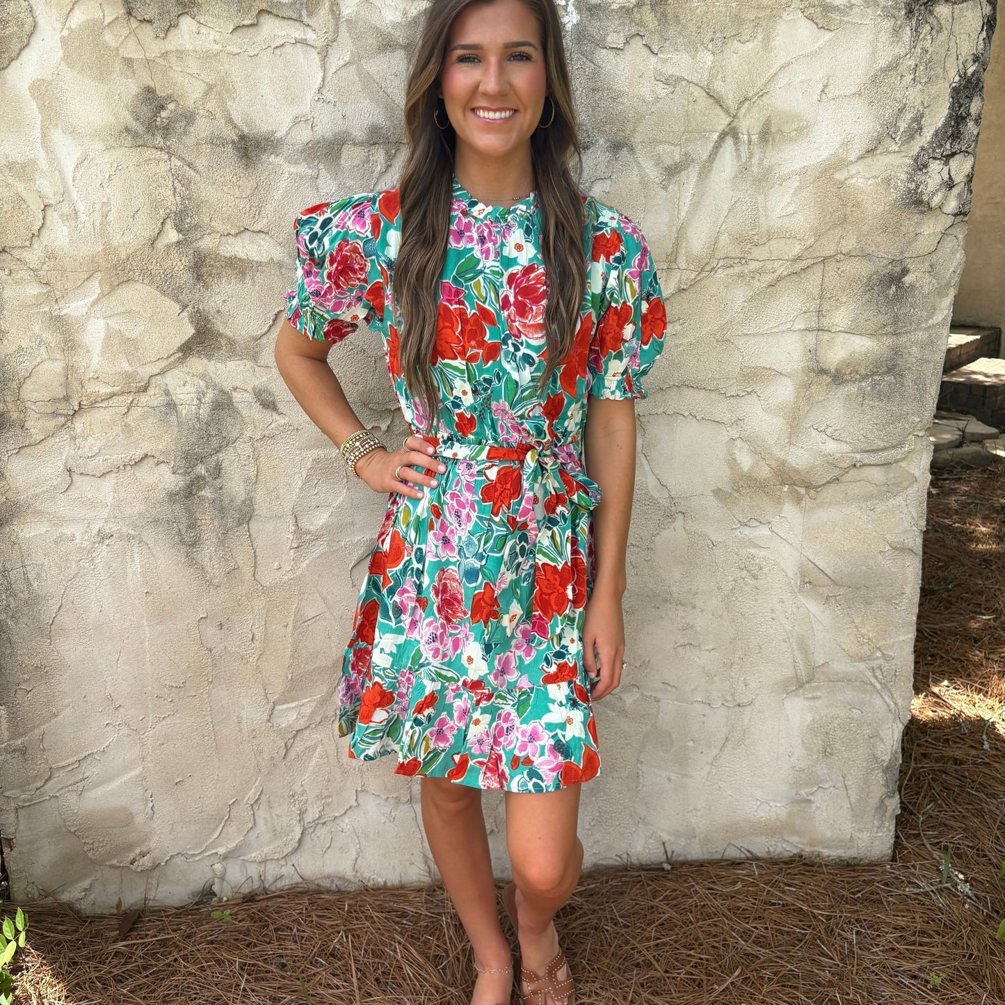 Woman wearing a colorful floral dress standing against a stone wall.