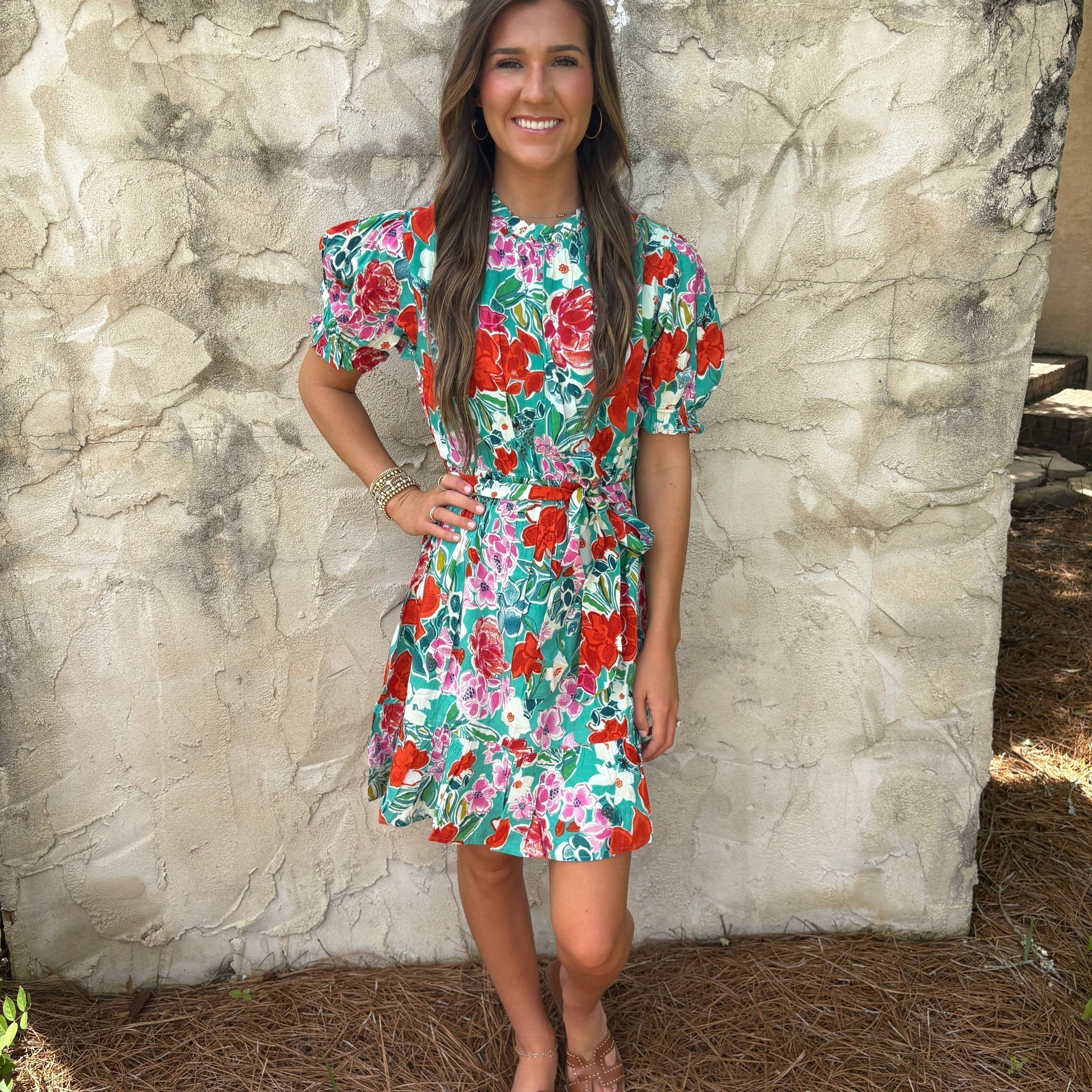 Woman wearing a colorful floral dress standing against a stone wall.