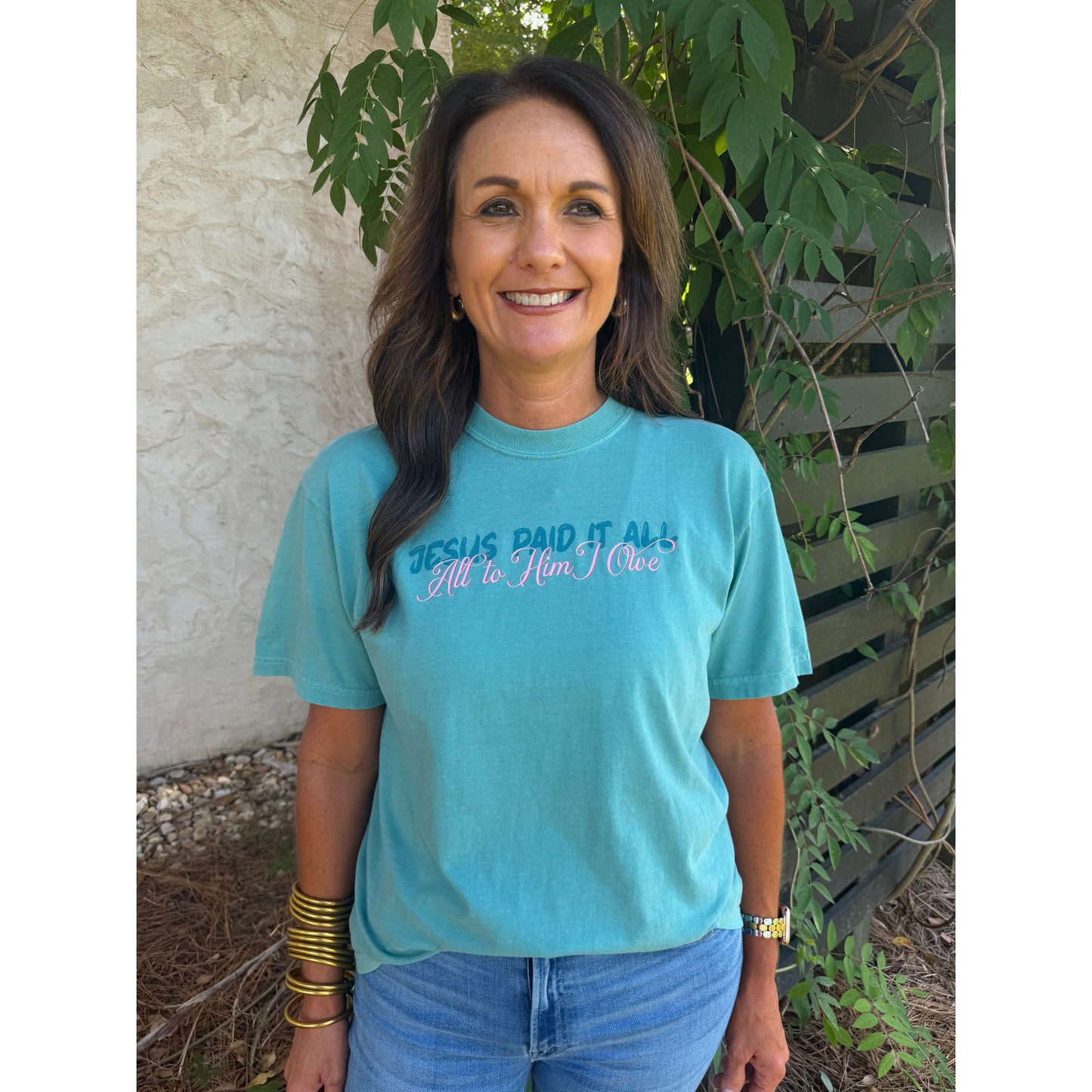 Woman wearing a turquoise t-shirt with text, standing outdoors against a wall with greenery.