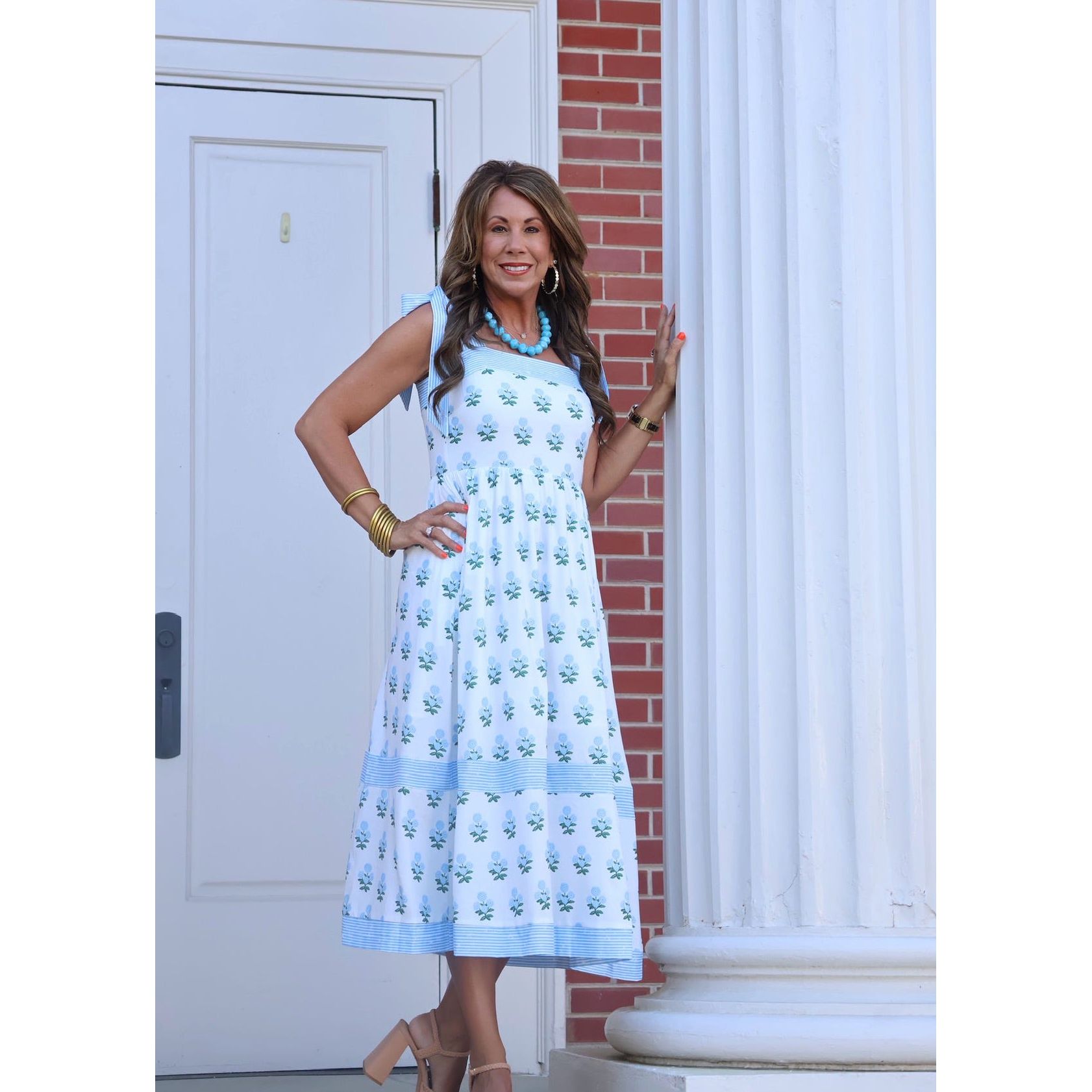 Woman in a light blue dress with floral pattern standing in front of a white door and brick wall.