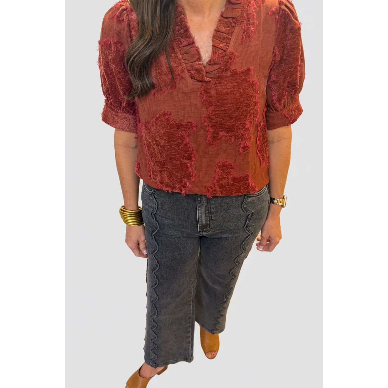 A woman wearing a rust red textured blouse with puff sleeves and dark gray scalloped wide-leg jeans, standing indoors near a white column.