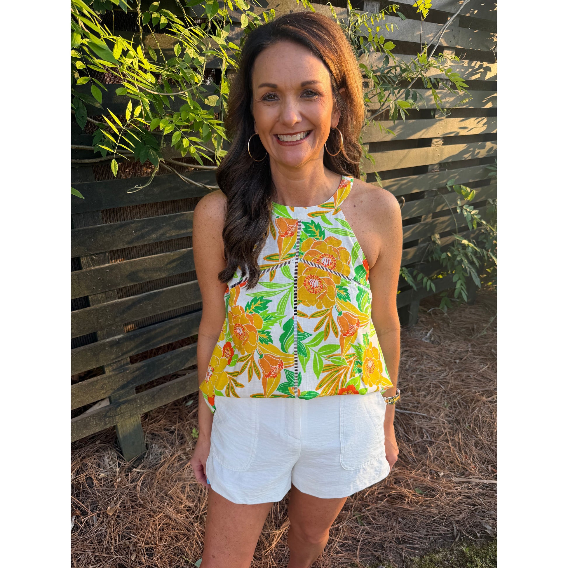 Woman wearing a colorful floral top and white shorts standing outdoors with greenery in the background