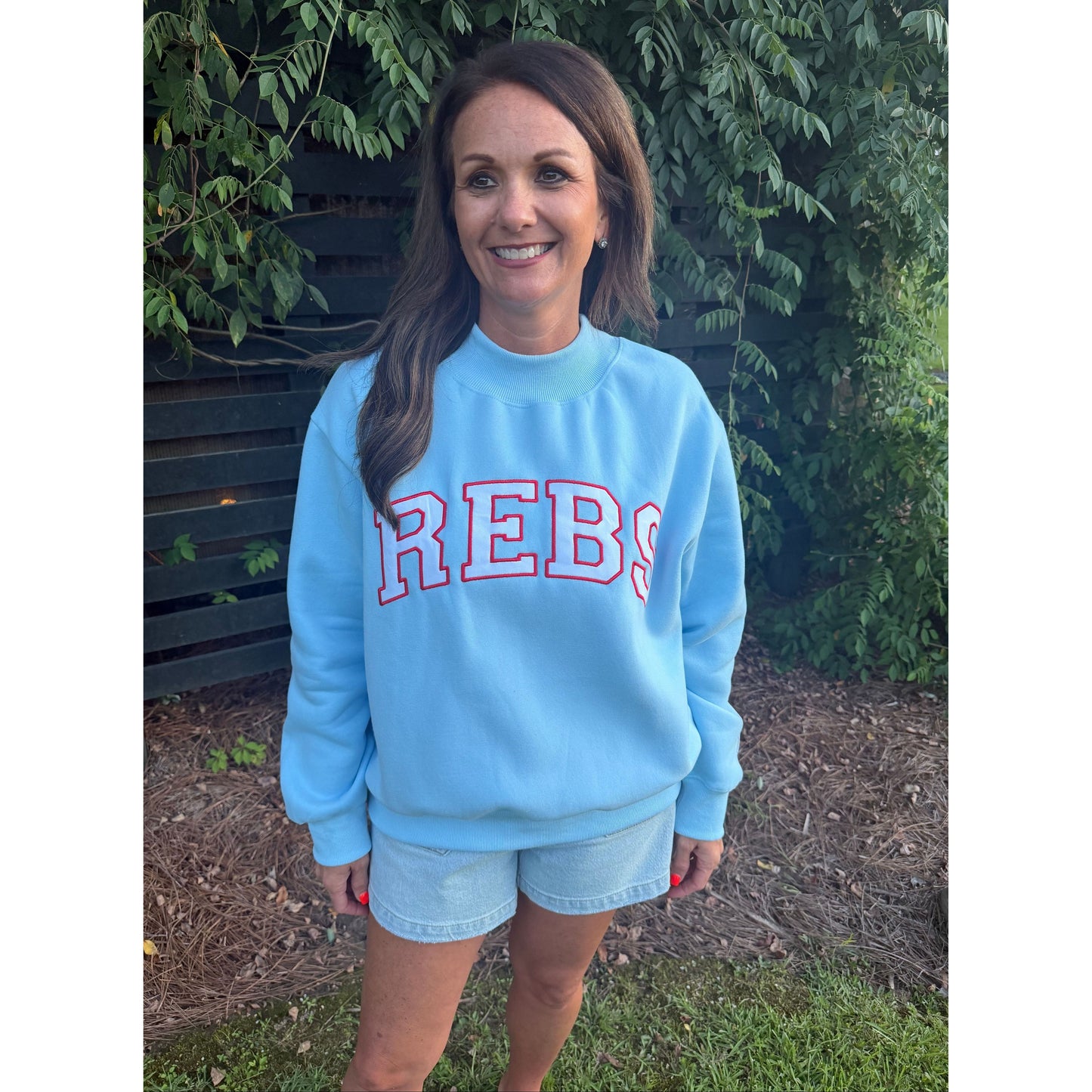 A woman wearing a light blue crewneck sweatshirt with 'REBS' printed in red and white block letters, paired with denim shorts, standing outdoors on a bench with green foliage and mulch in the background.