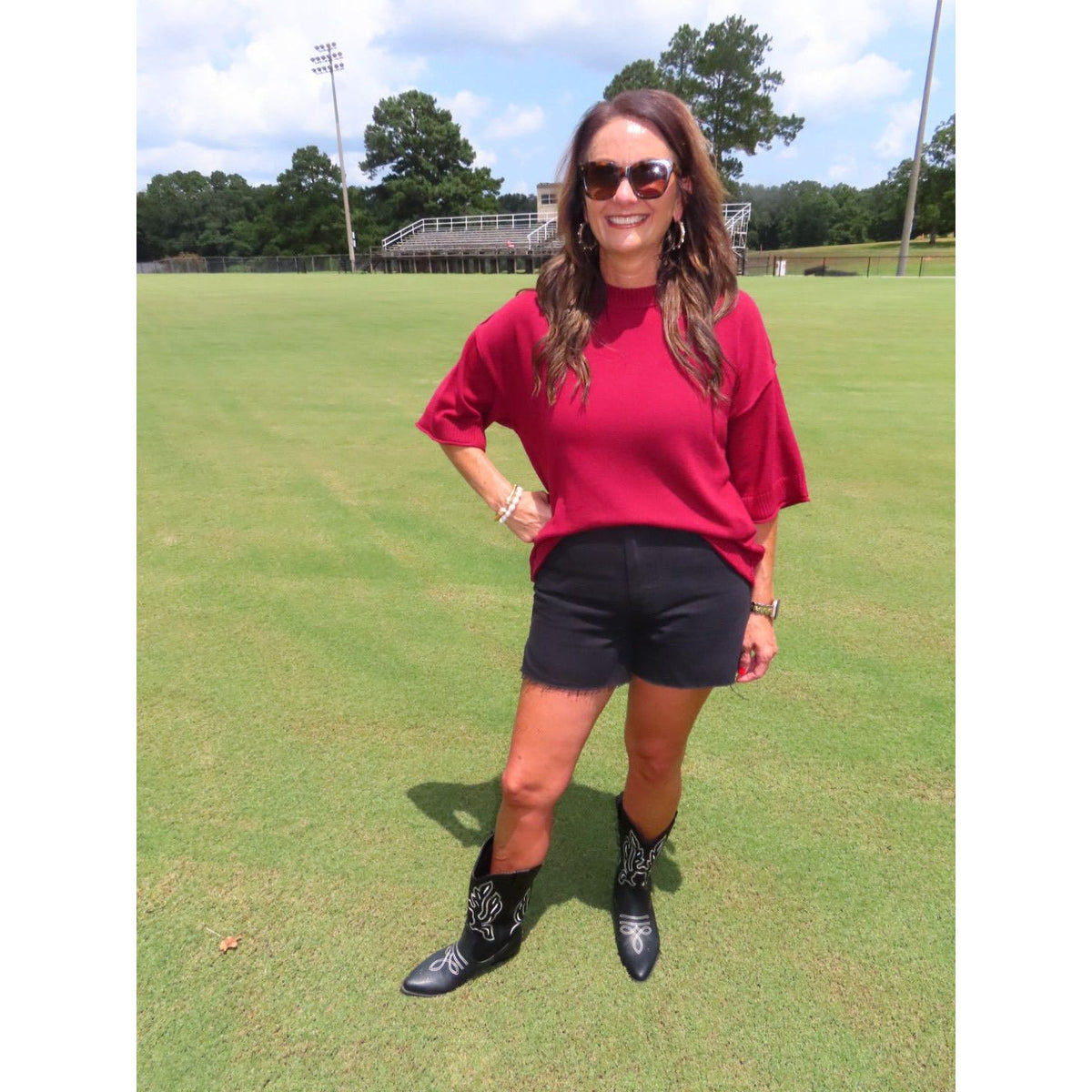 A woman stands on a green grass field wearing a bright red short-sleeve t-shirt, black denim shorts, and black western-style boots with silver embroidery. She is also wearing sunglasses and has her hand on her hip.