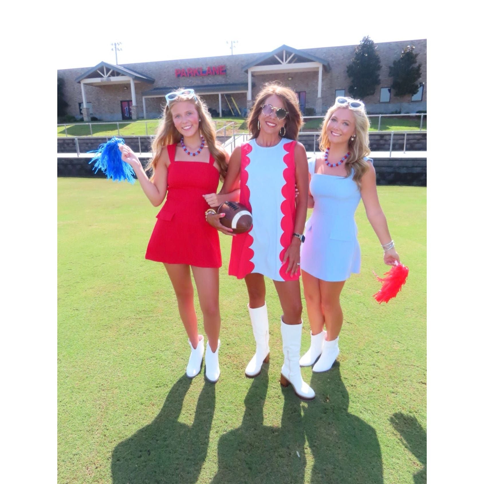 Three women posing together on a green grass field wearing matching retro-style mini dresses in red, white, and light blue, holding pom-poms and a football-shaped bag.