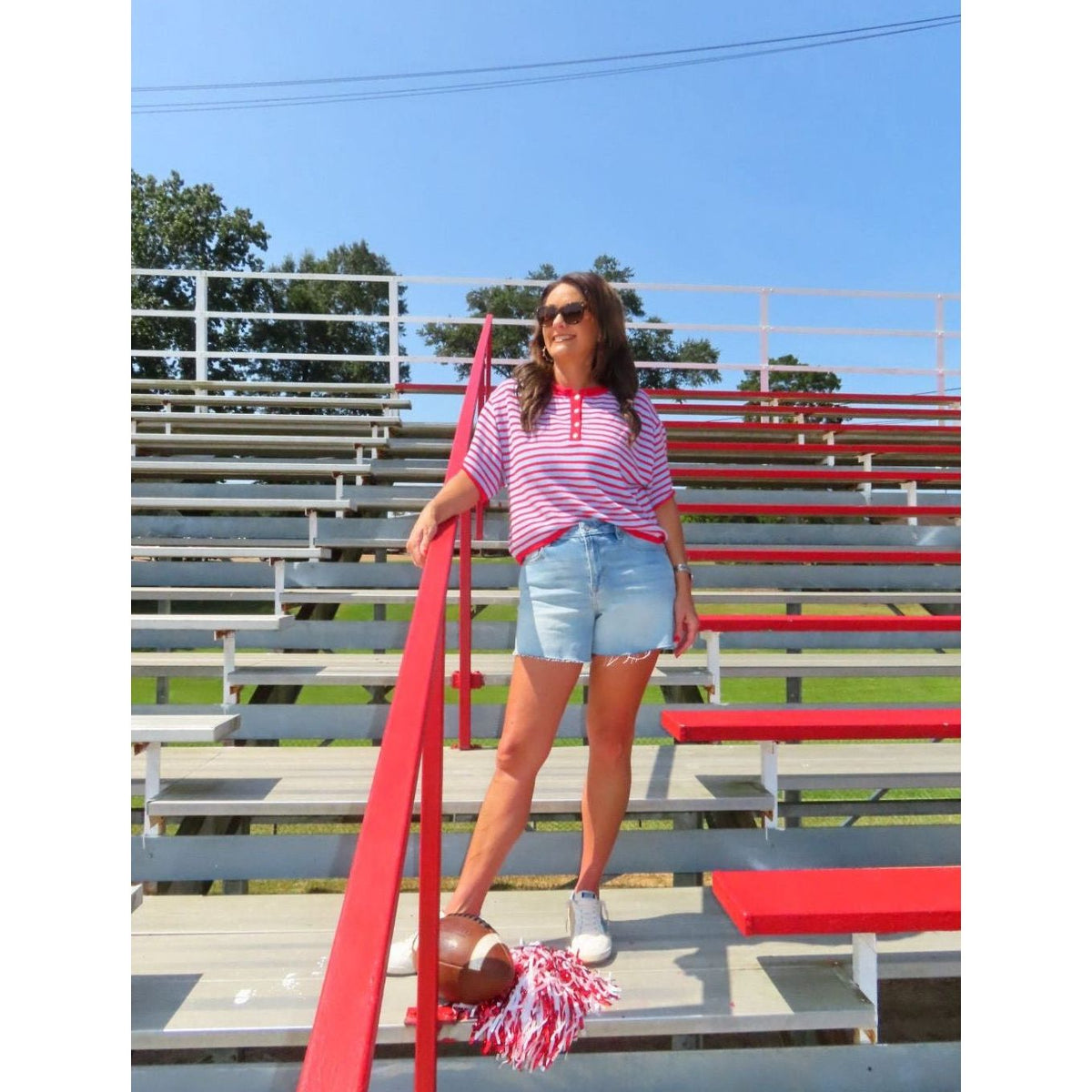 A woman stands on red and gray outdoor stadium bleachers under a blue sky, wearing a red and white striped short-sleeve polo shirt, light blue denim shorts, white sneakers, and holding a red and white pom-pom.