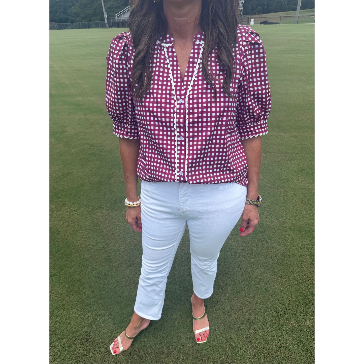 A woman wearing a burgundy and white gingham short-sleeve blouse, white pants, and white heeled sandals, standing on a green grassy field.