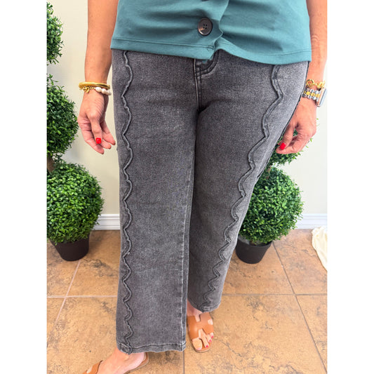 A person wearing gray denim jeans with scalloped seams down the sides, paired with a teal top and tan sandals, standing indoors near potted green plants.