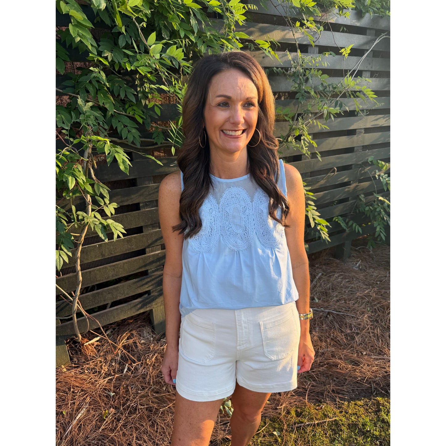 Woman wearing a light blue top and white shorts standing outdoors with greenery in the background