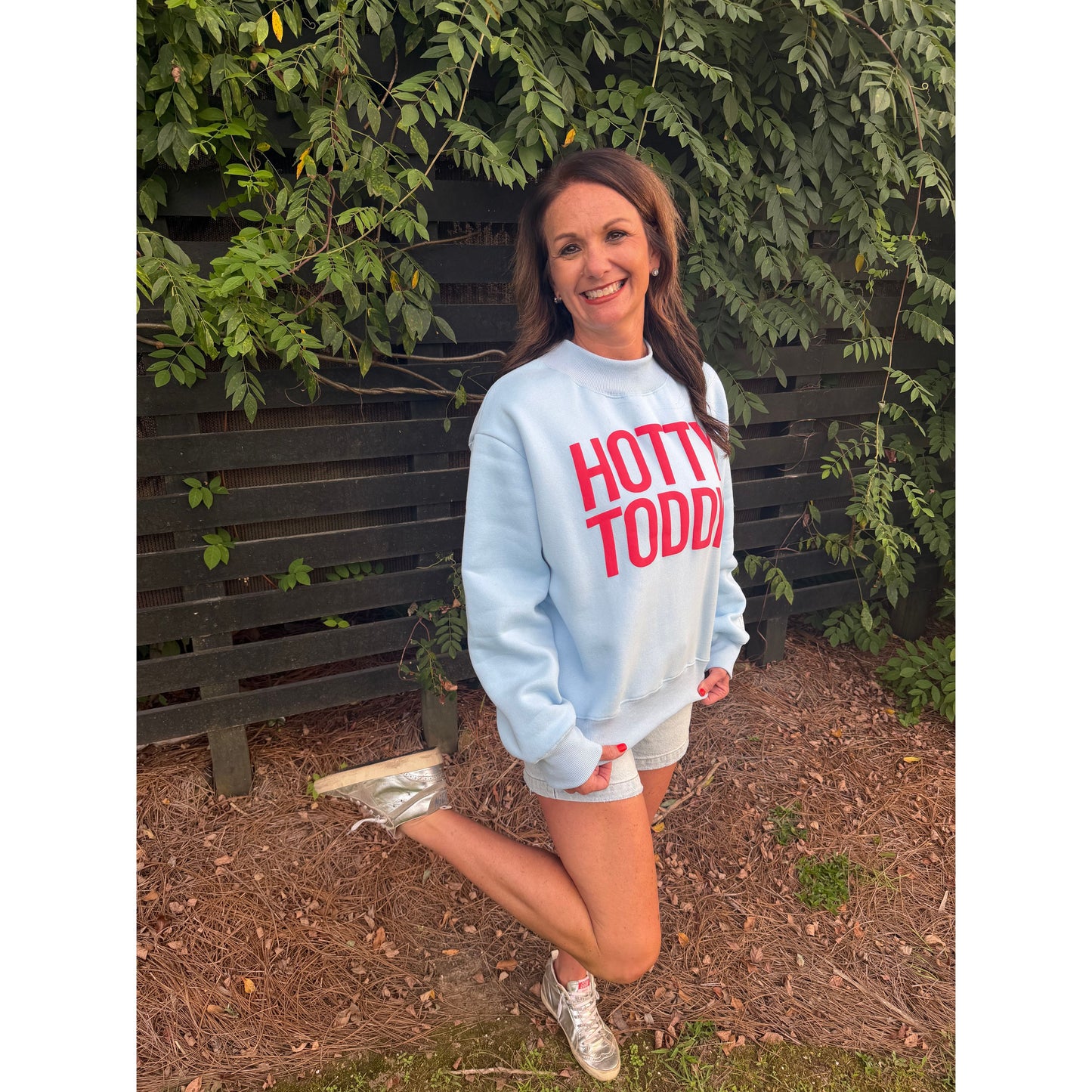 A woman posing outdoors in front of a wooden fence and green foliage, wearing a light blue sweatshirt with 'HOTTI TODDI' printed in red across the chest, paired with white shorts and sneakers.