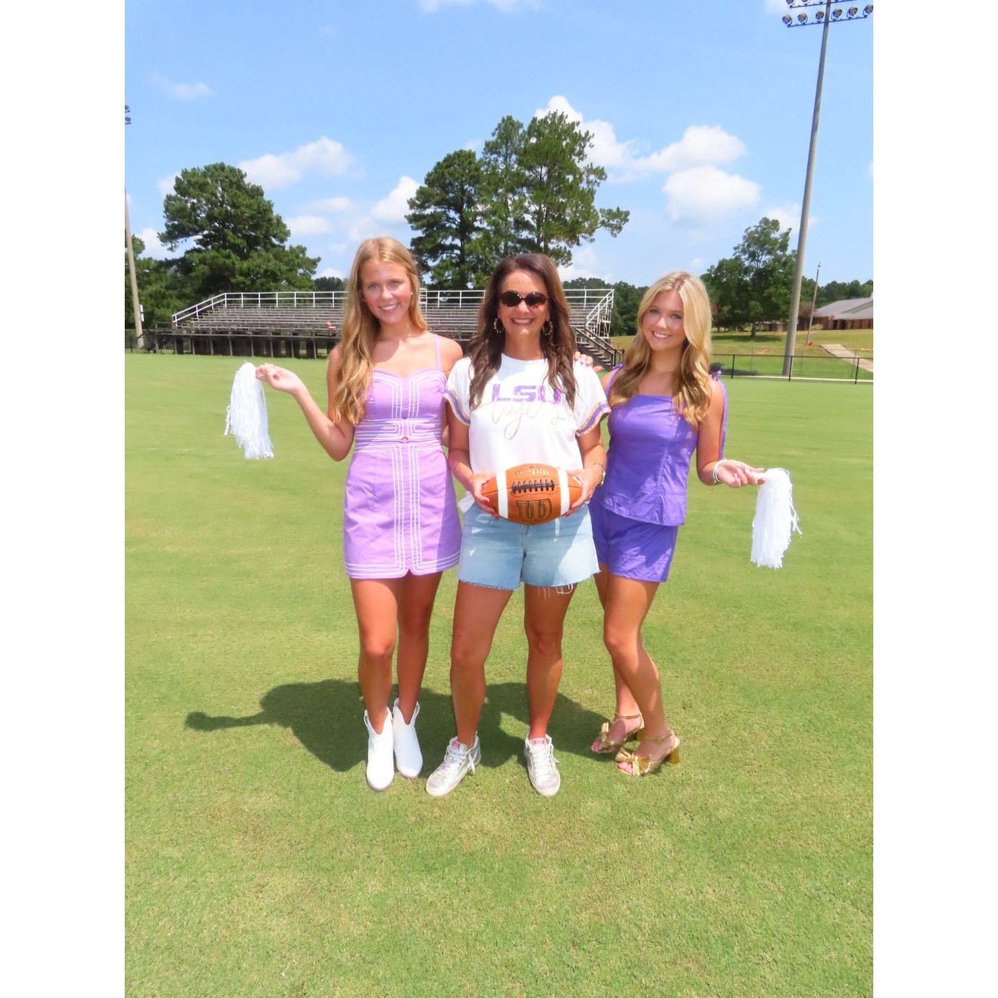 Three women standing on a grassy field under a blue sky, wearing coordinated purple and white athletic outfits and holding white pom-poms and an orange football.