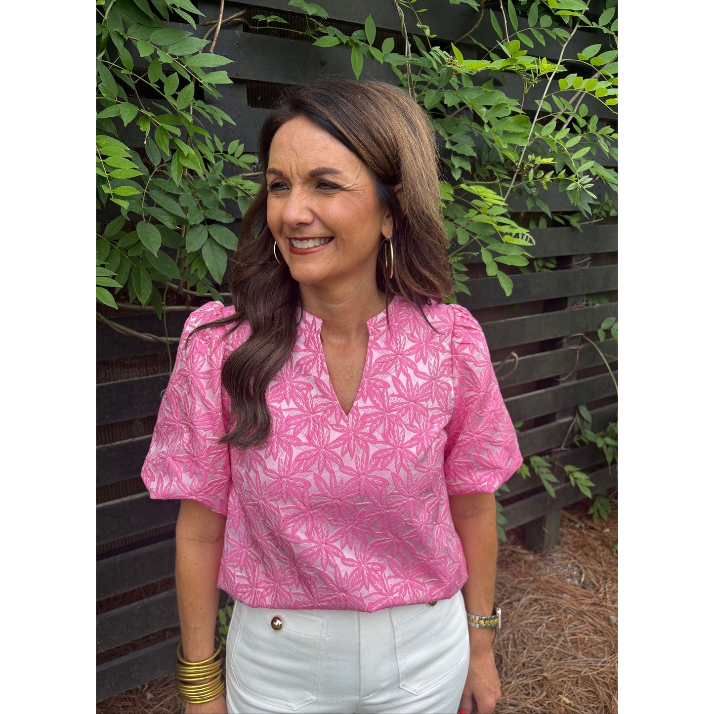 Woman wearing a pink patterned shirt and white pants sitting on a bench with greenery around.