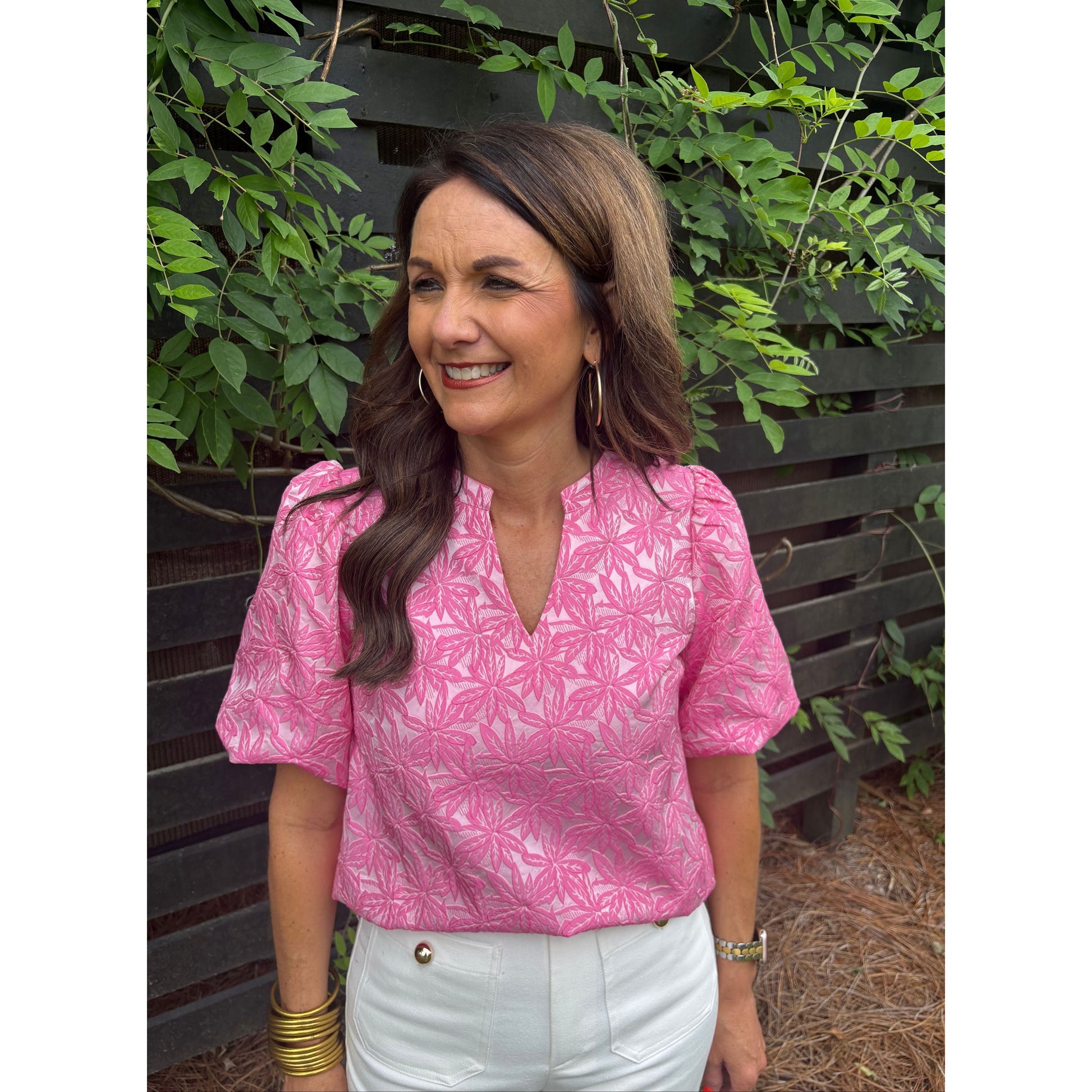 Woman wearing a pink patterned shirt and white pants sitting on a bench with greenery around.