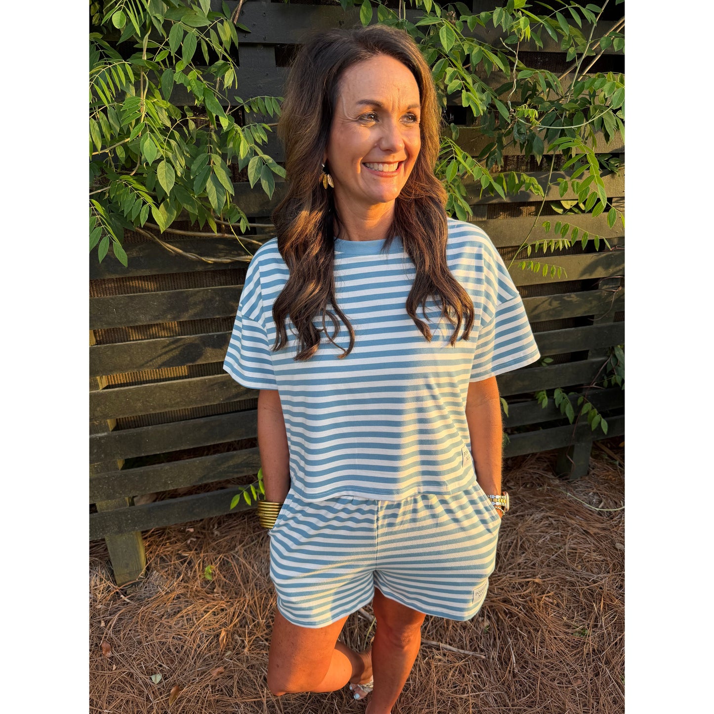 Woman wearing a blue and white striped dress standing outdoors with greenery and a wooden bench in the background.