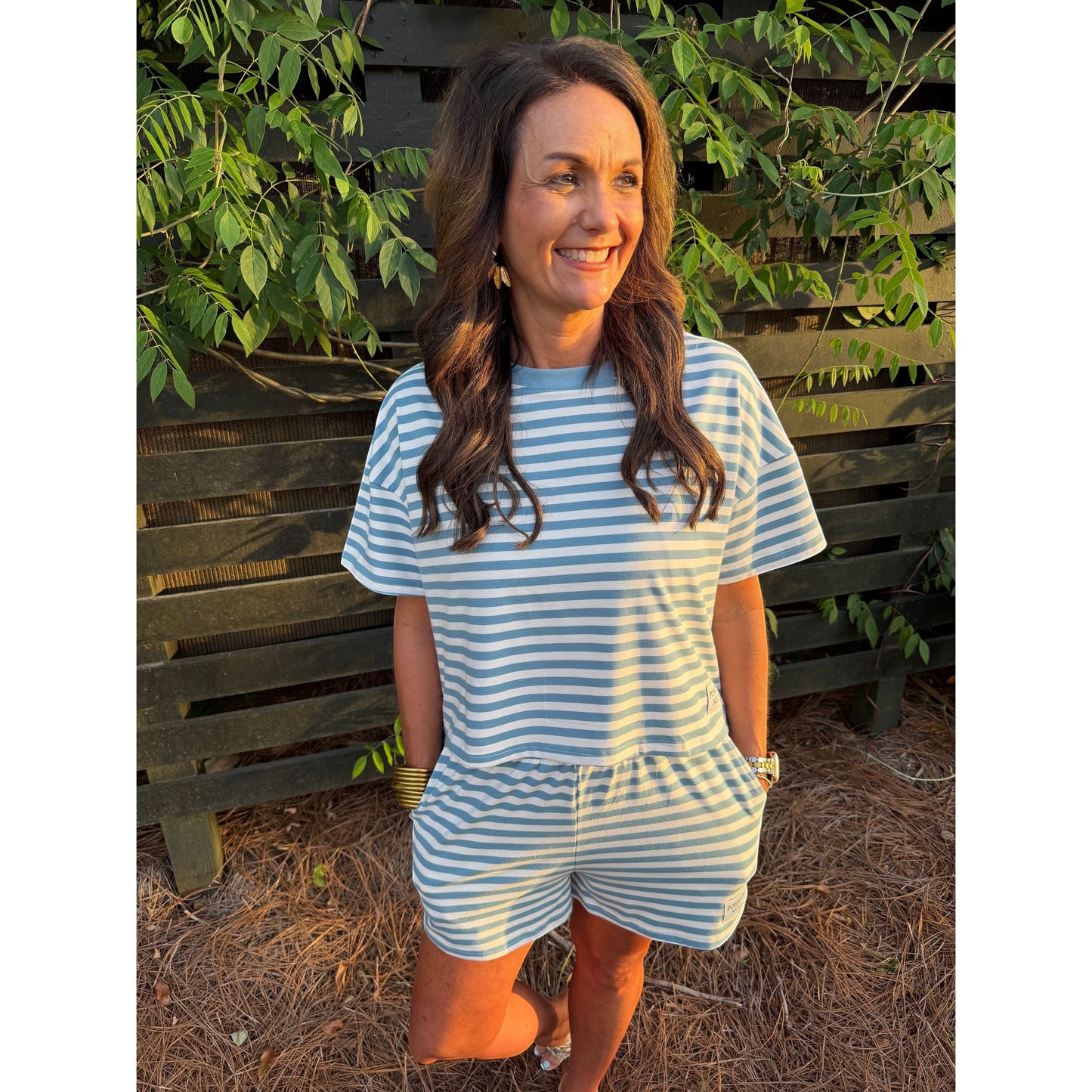 Woman wearing a blue and white striped dress standing outdoors with greenery and a wooden bench in the background.