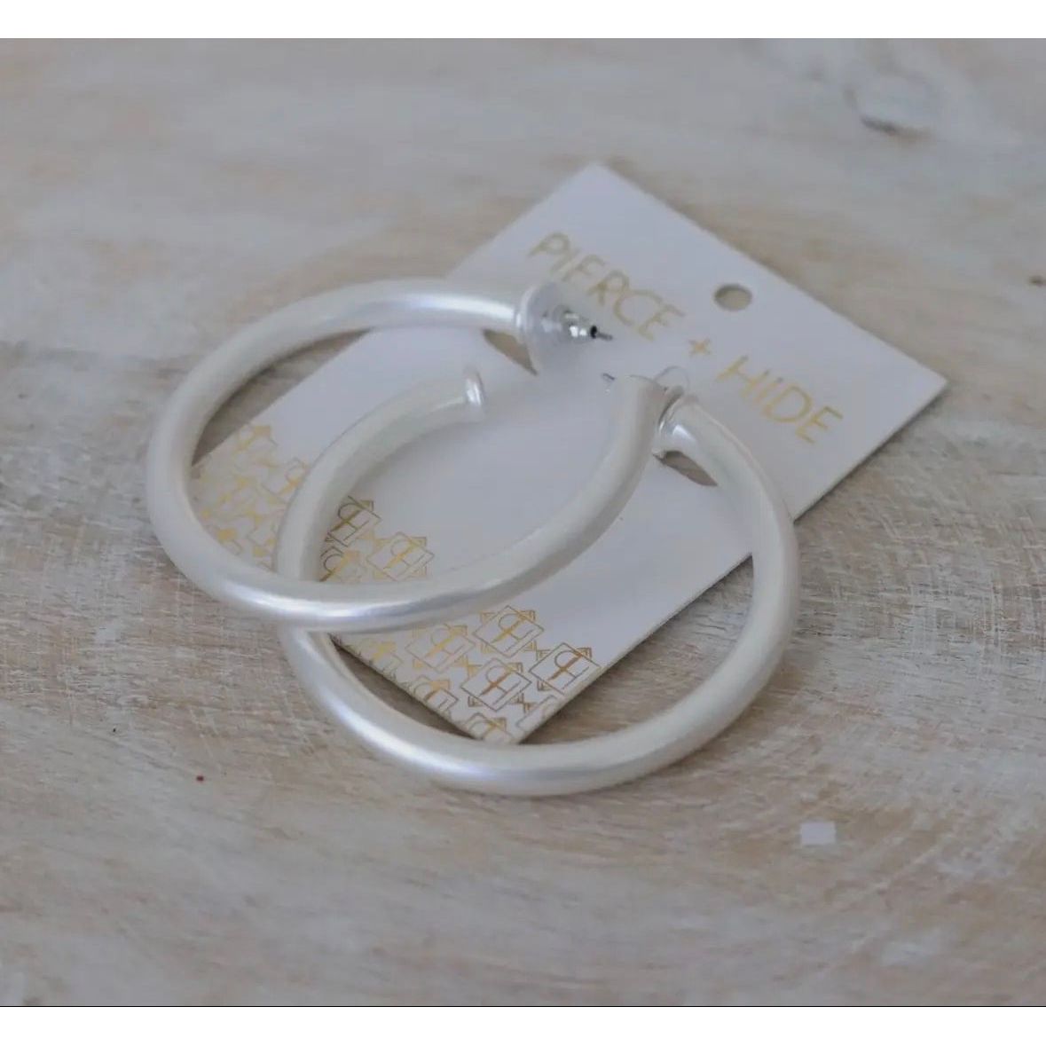 Silver hoop earrings on a beige card with 'Pierce + Hide' branding on a wooden surface.