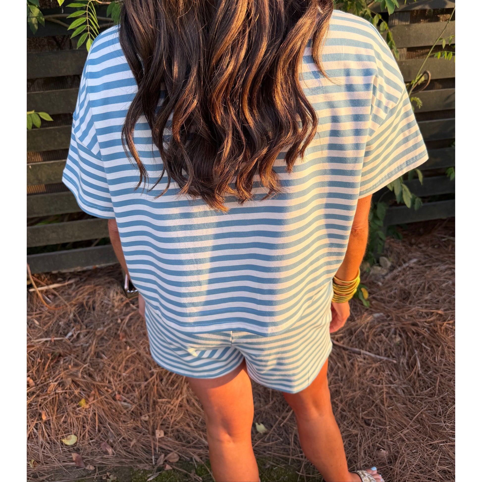 Person wearing a blue and white striped shirt and shorts standing outdoors with greenery in the background.