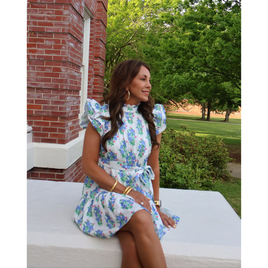 Woman in a floral dress standing outdoors with trees and a brick building in the background
