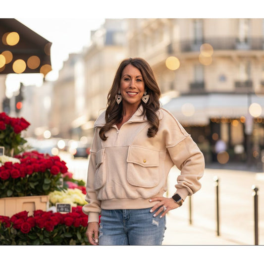 Woman wearing a beige sweater and blue jeans standing against a light gray brick wall.
