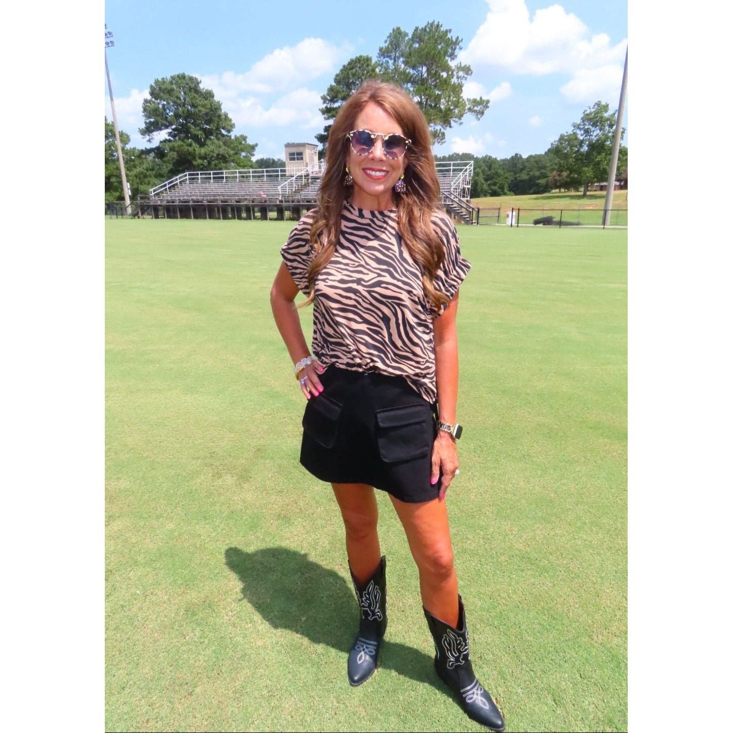 A woman stands on a grassy field wearing a brown and black zebra-print short-sleeve top, a black mini skirt, and black western boots with silver embroidery. She is wearing sunglasses and smiling.