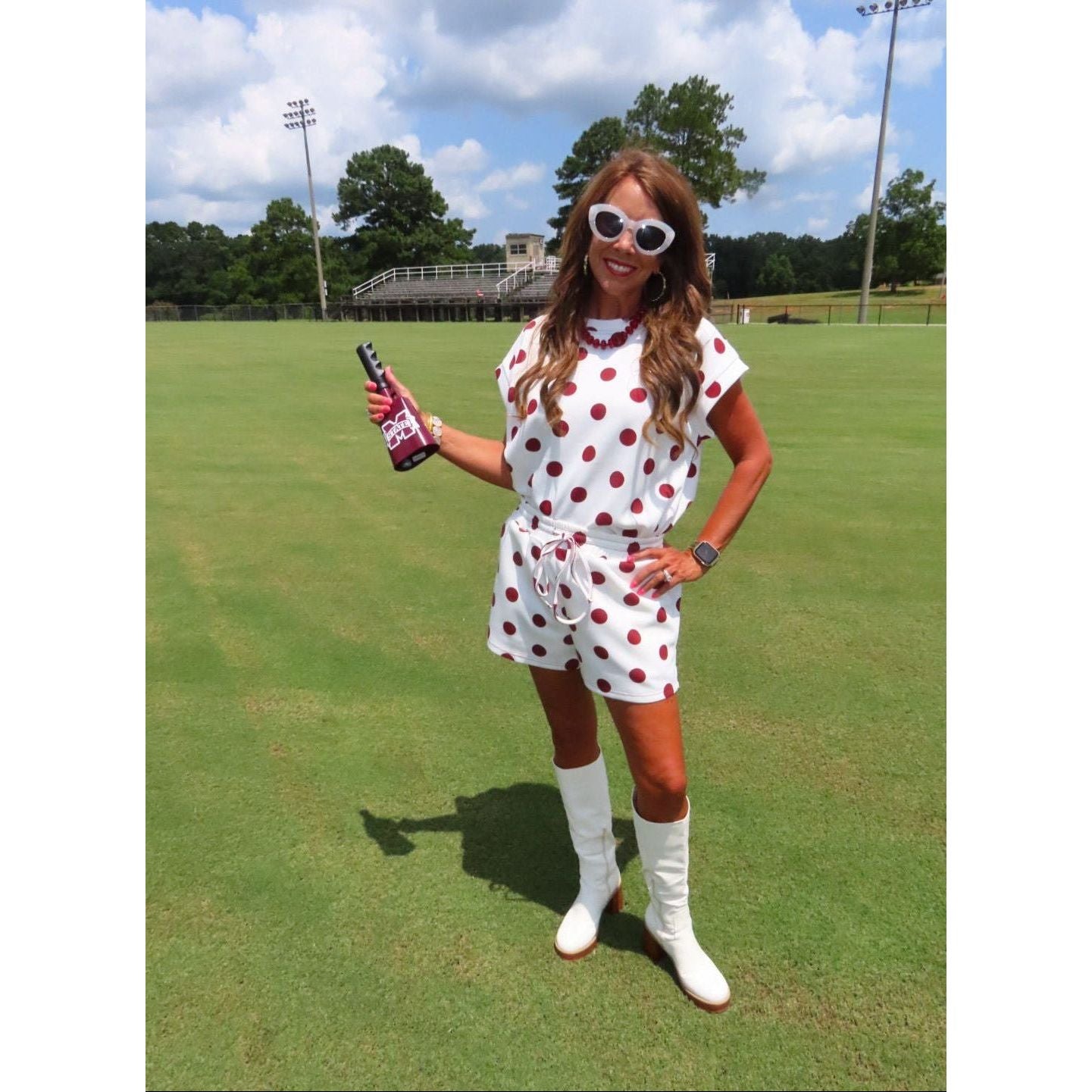 A woman stands on a green grass field wearing a white short-sleeve romper and matching white knee-high socks with red polka dots, white knee-high boots, and white-framed sunglasses while holding a bottle.
