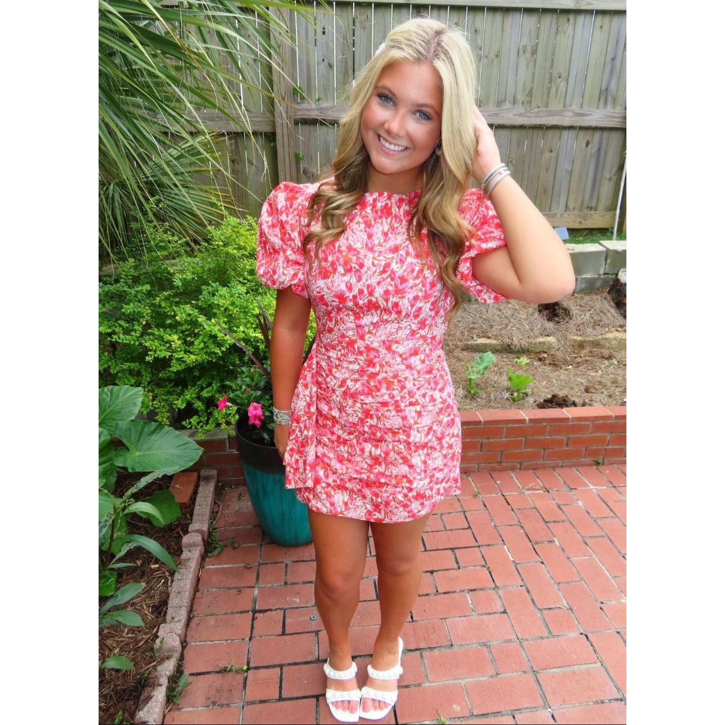 Woman in a red floral dress standing outdoors on a brick path with plants and a wooden fence in the background.