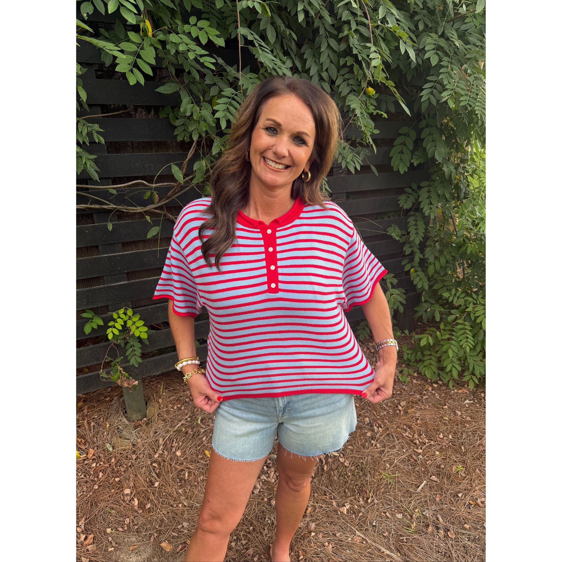 A woman wearing a red and white striped short-sleeve henley top with light blue denim shorts, standing outdoors in front of a dark wooden fence and green foliage.