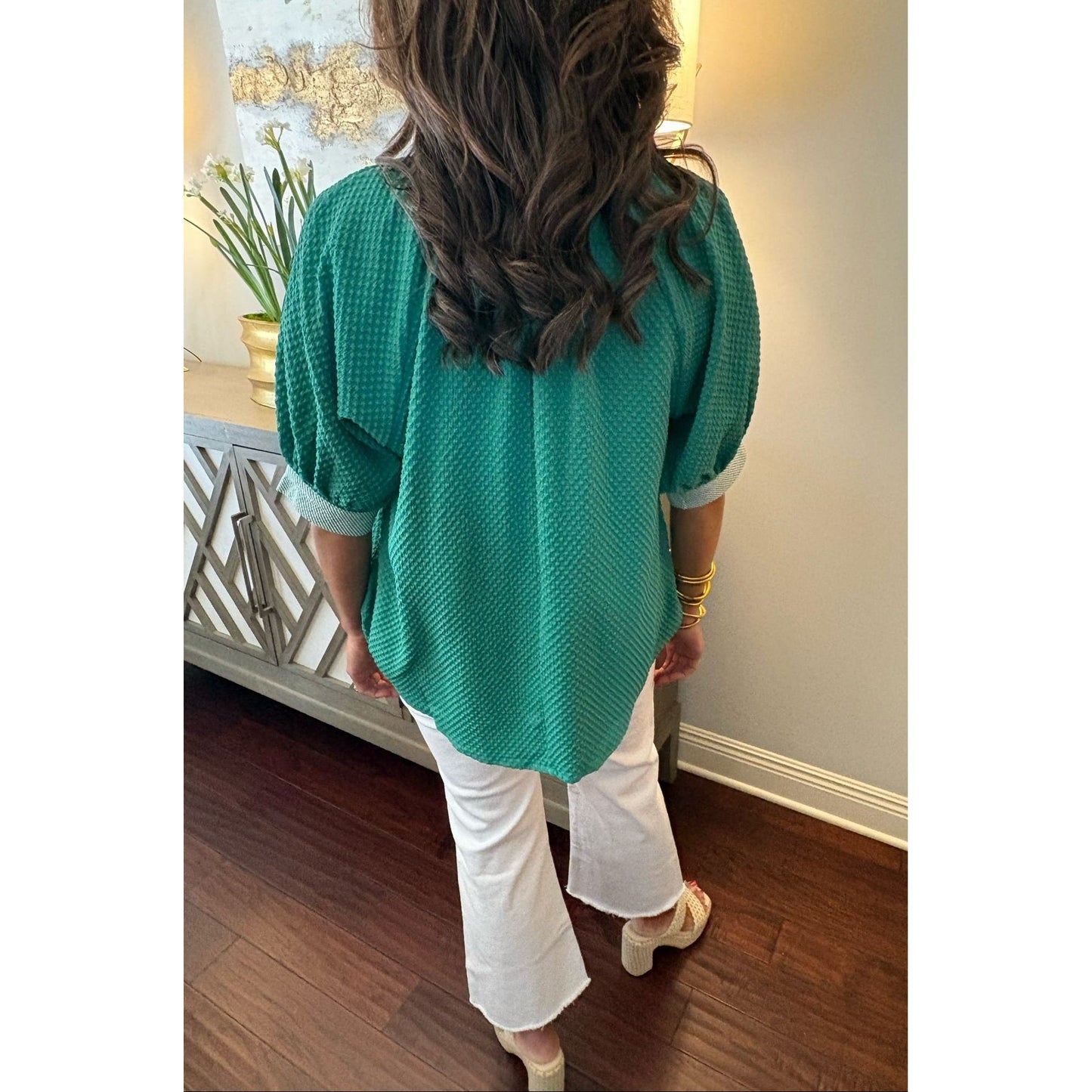 Person wearing a green sweater and white pants in a room with wooden flooring and a decorative cabinet.
