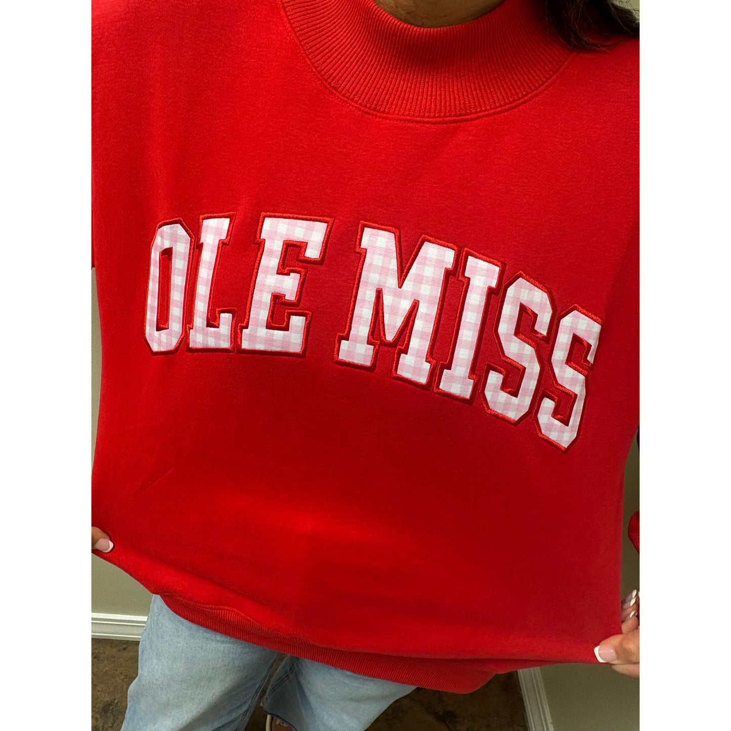 A close-up of a person wearing a bright red crewneck sweatshirt with 'OLE MISS' in white gingham lettering outlined in red across the chest, paired with light blue jeans.