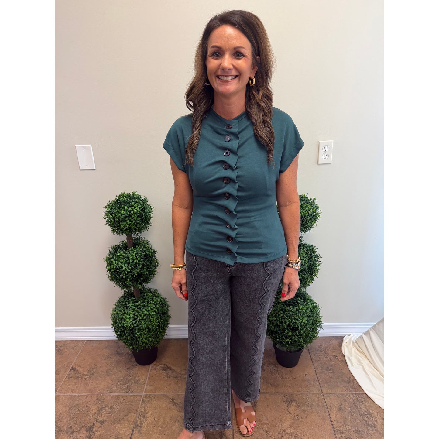 A woman modeling a short-sleeve teal button-front top and dark gray patterned pants, standing indoors between two potted topiary trees against a beige wall.