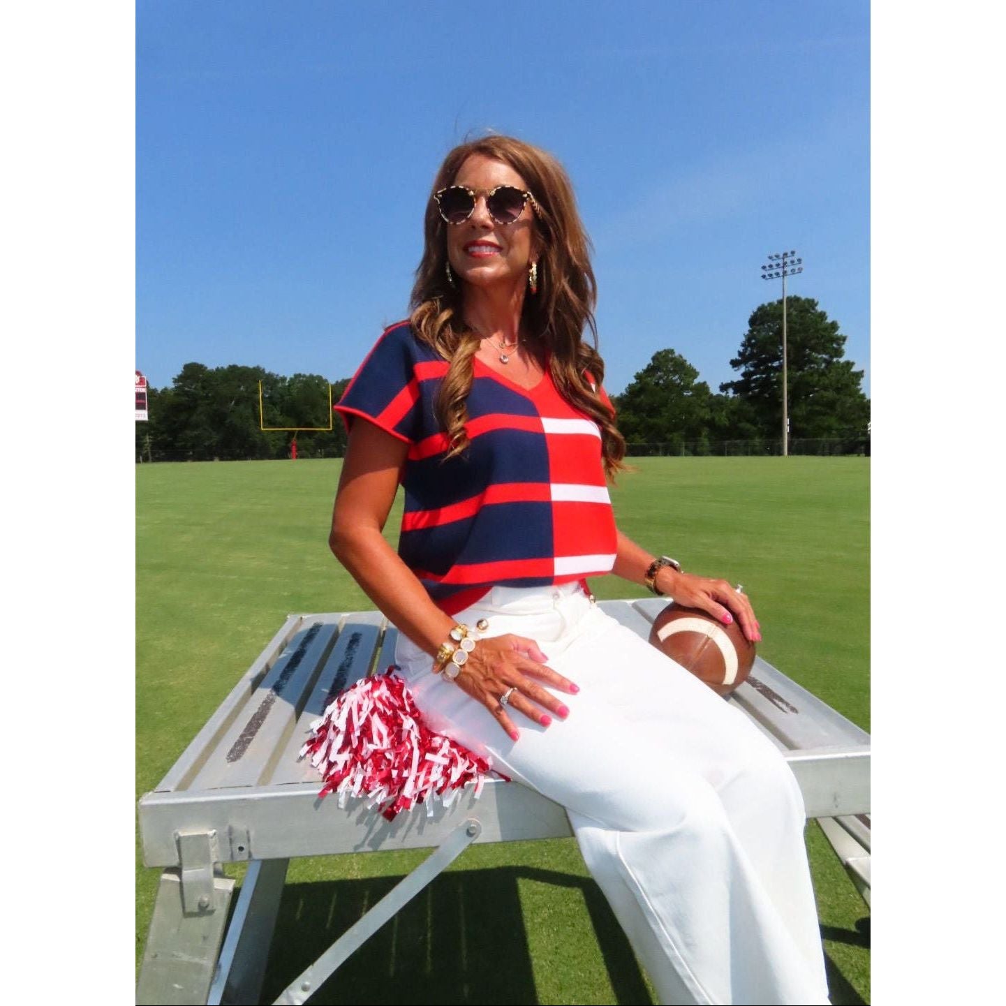 A woman wearing a red, navy, and white striped short-sleeve crop top and white pants sits on a metal bleacher at a grassy field under a blue sky, holding a football and red/white pom-poms.
