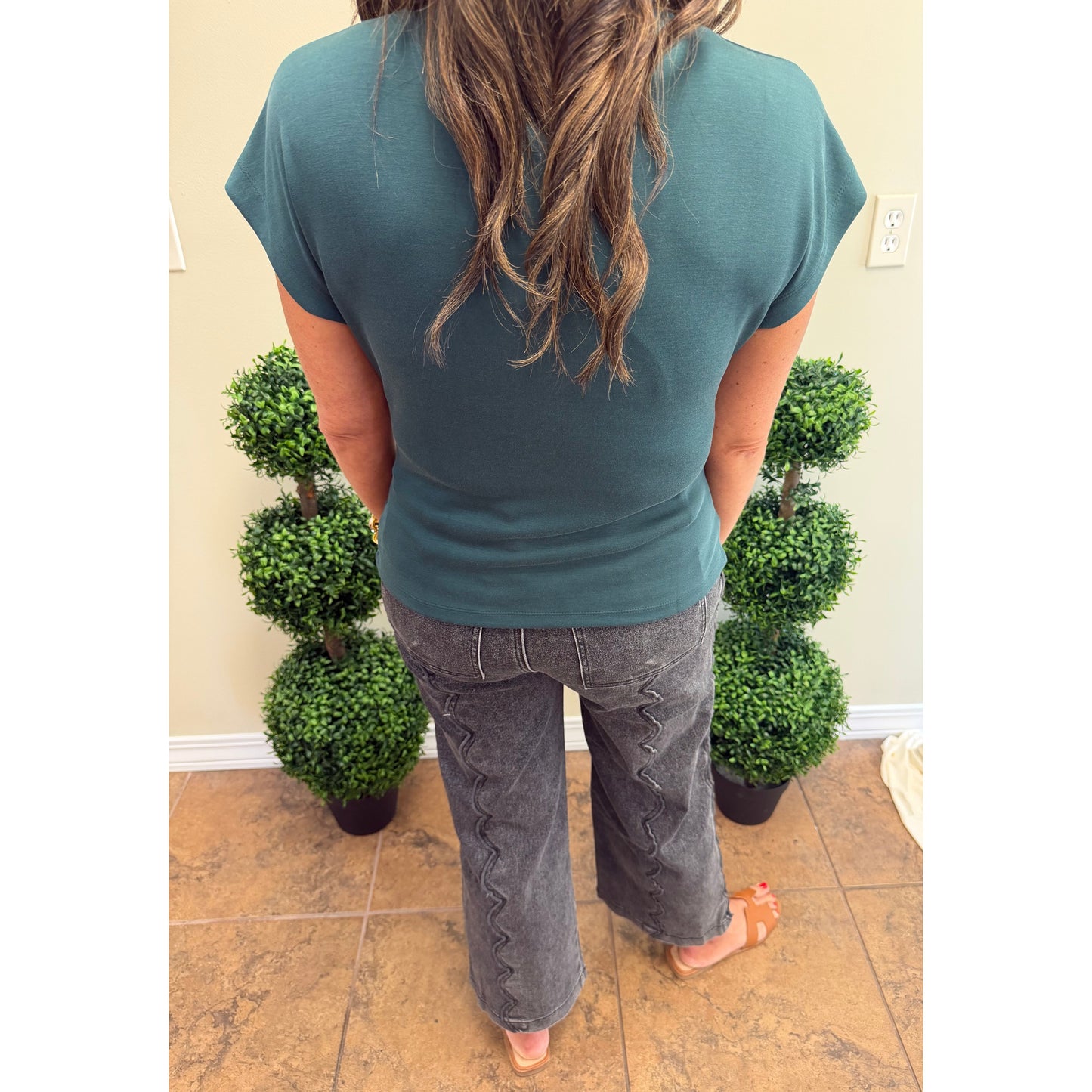 A woman viewed from behind wearing a teal short-sleeve t-shirt and gray wide-leg jeans with decorative zigzag stitching down the back, standing indoors between two potted topiary trees.