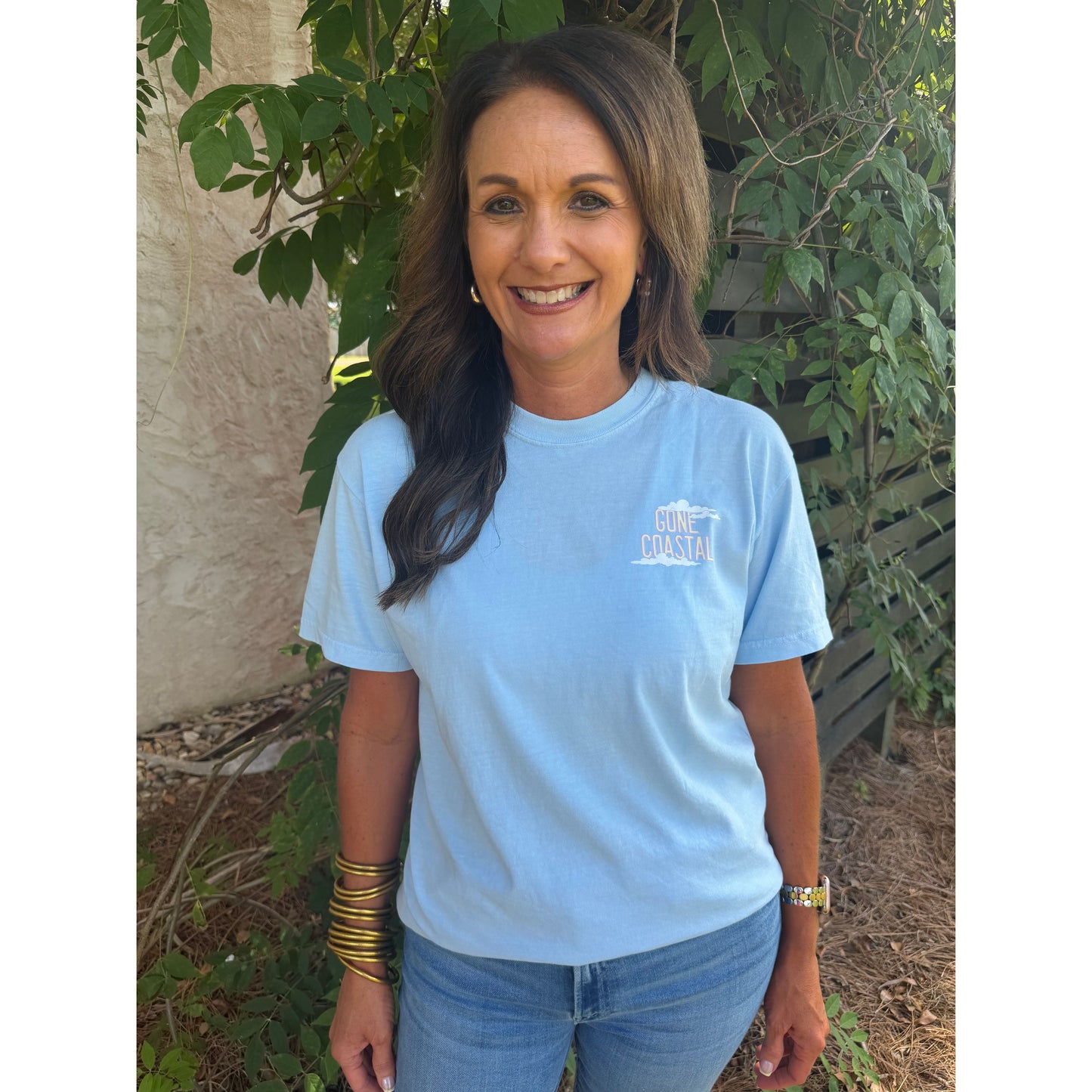 Woman wearing a light blue t-shirt with text, standing outdoors near greenery.
