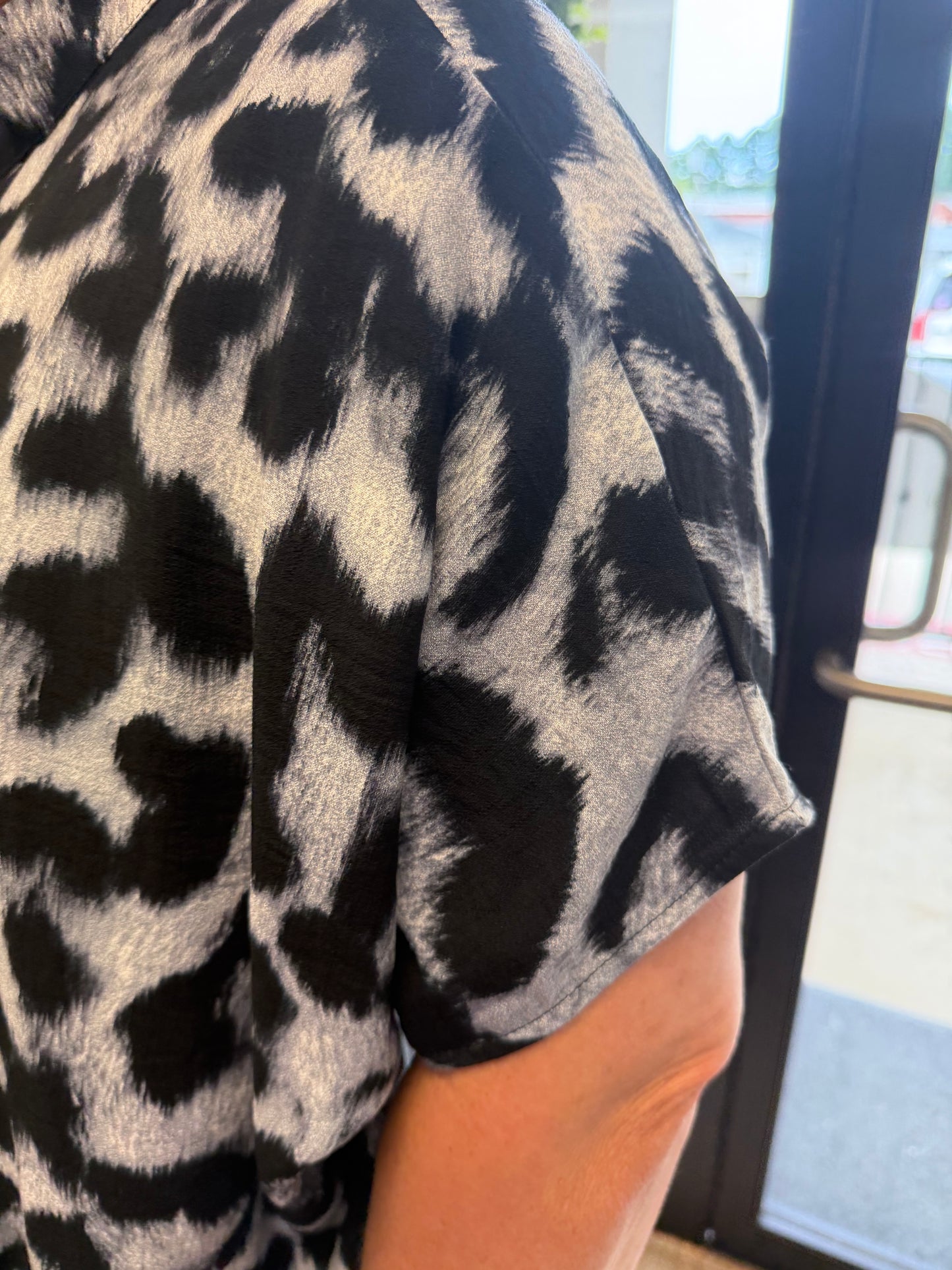 Close-up of a person wearing a short-sleeve top with a black and gray leopard print pattern, standing indoors near a glass door.
