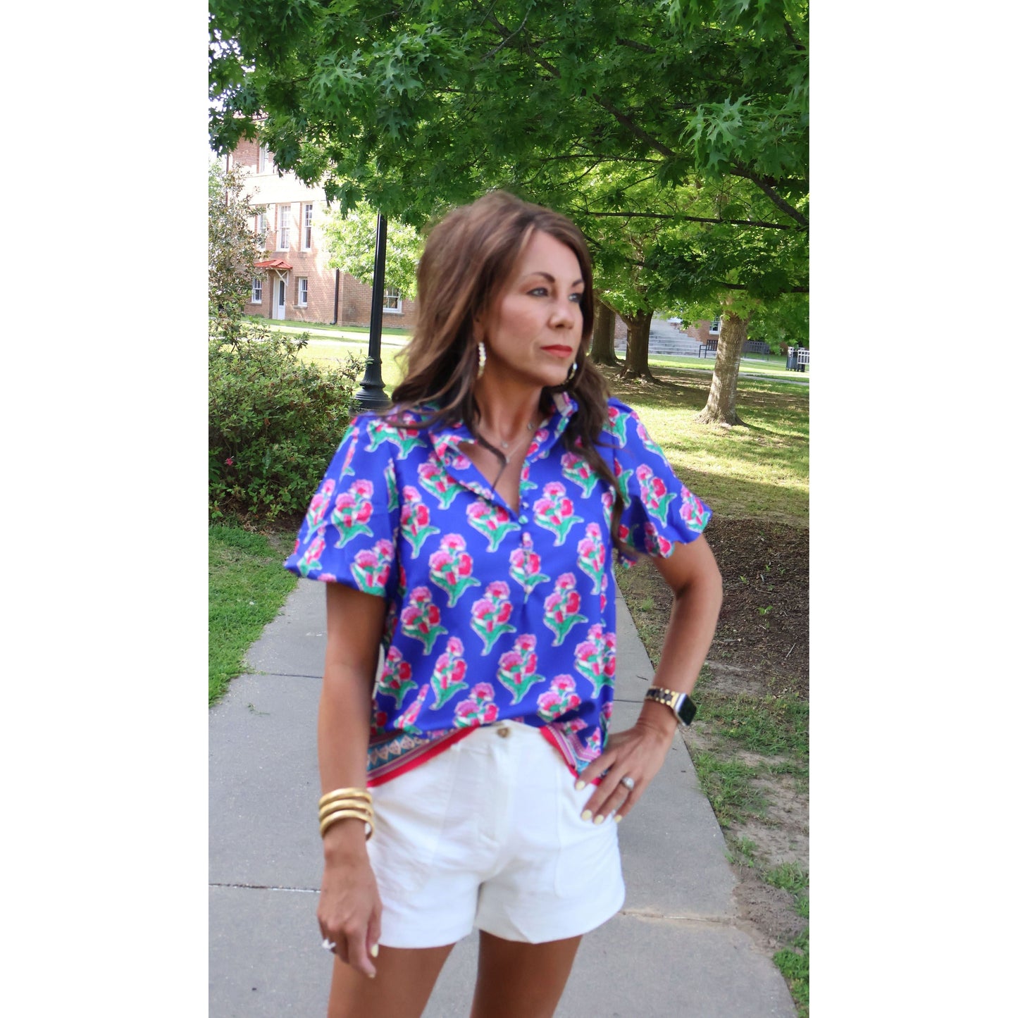 Woman wearing a colorful floral blouse and white shorts standing on a sidewalk with trees and grass in the background.