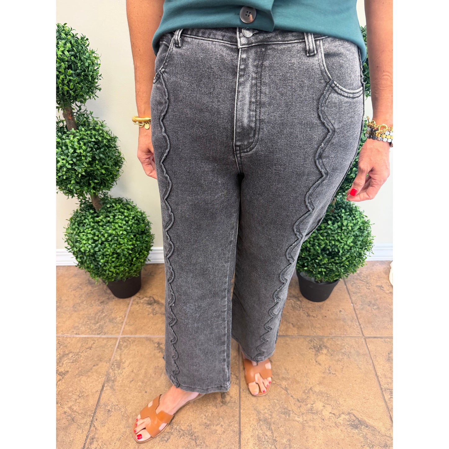A person wearing gray washed denim jeans with distinctive wavy seam detailing down the outer sides, paired with a teal top and tan slide sandals, standing on a tiled floor between potted green plants.