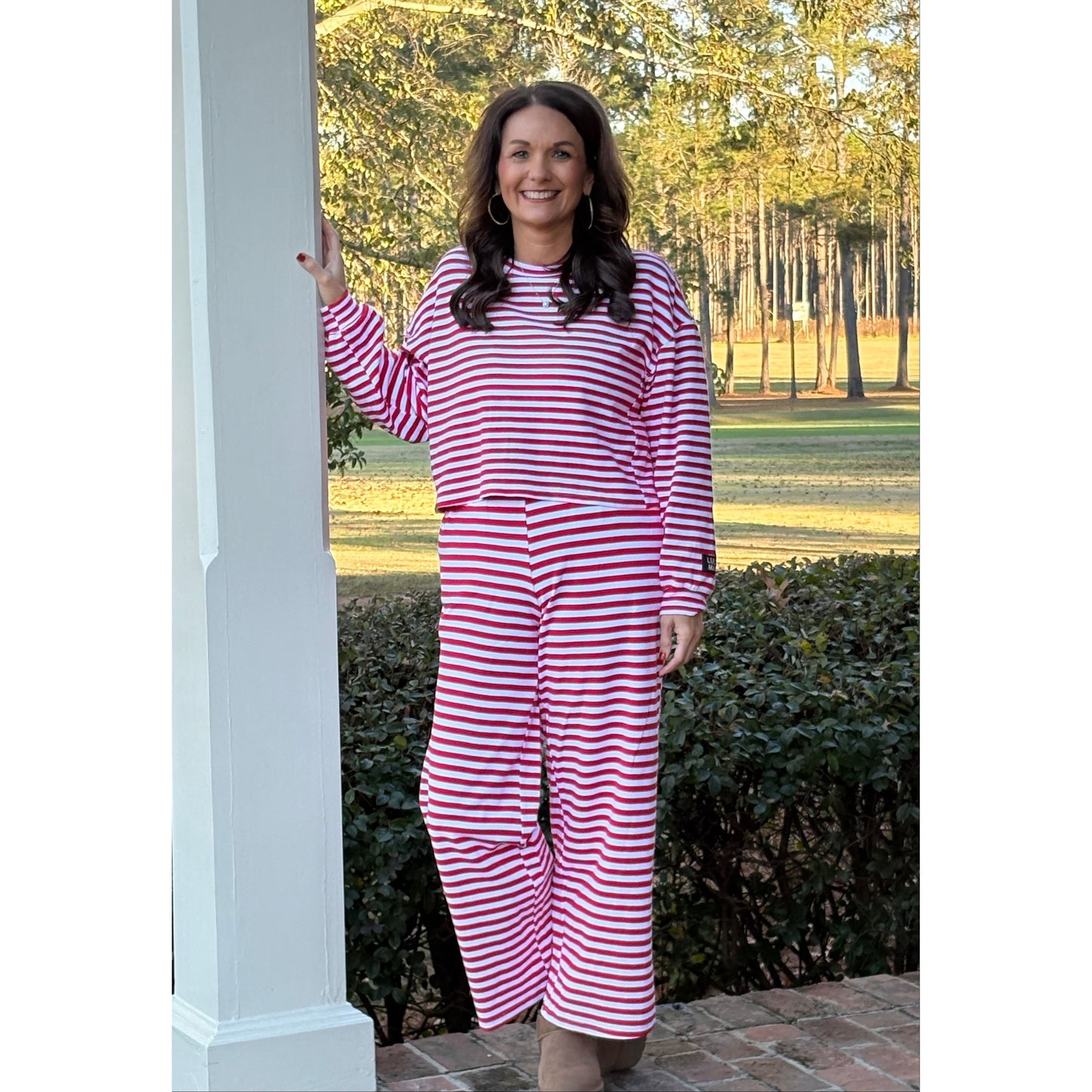 Woman wearing a red and white striped dress standing outdoors with trees and a brick wall in the background.