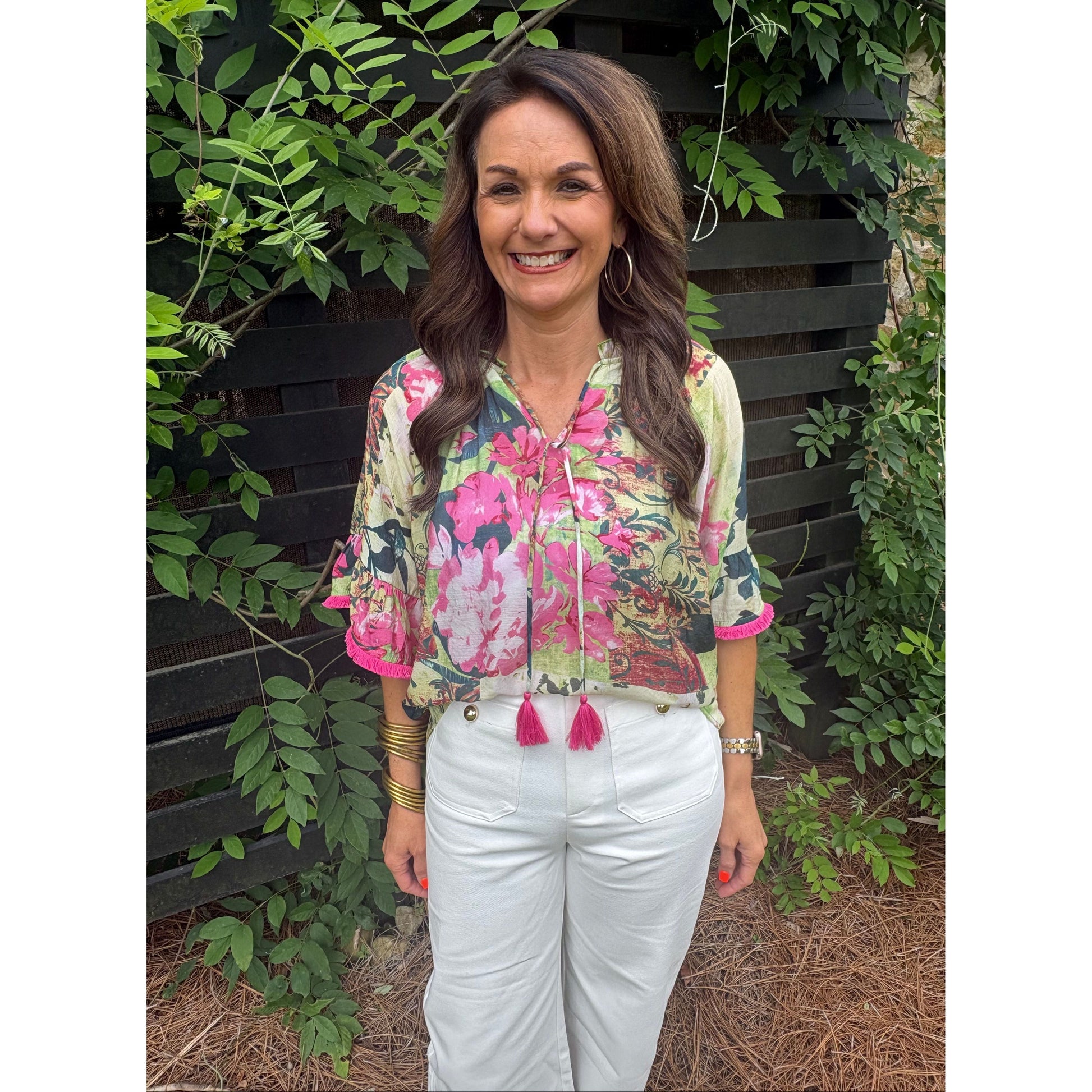 Woman wearing a colorful floral blouse with tassels standing in front of greenery.