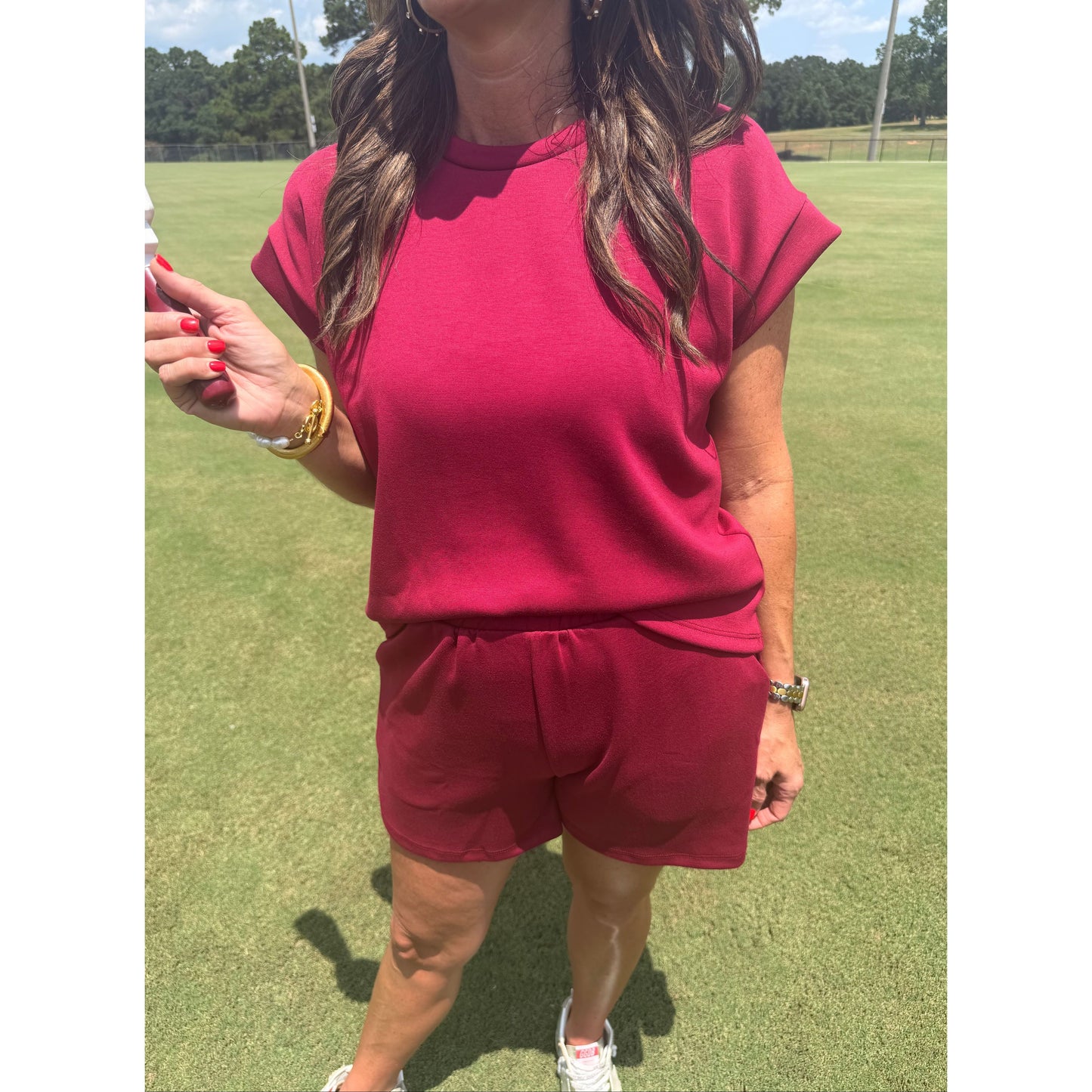 A woman wearing a matching two-piece outfit consisting of a short-sleeve crewneck top and shorts in a deep magenta pink, photographed outdoors on a green grass field.