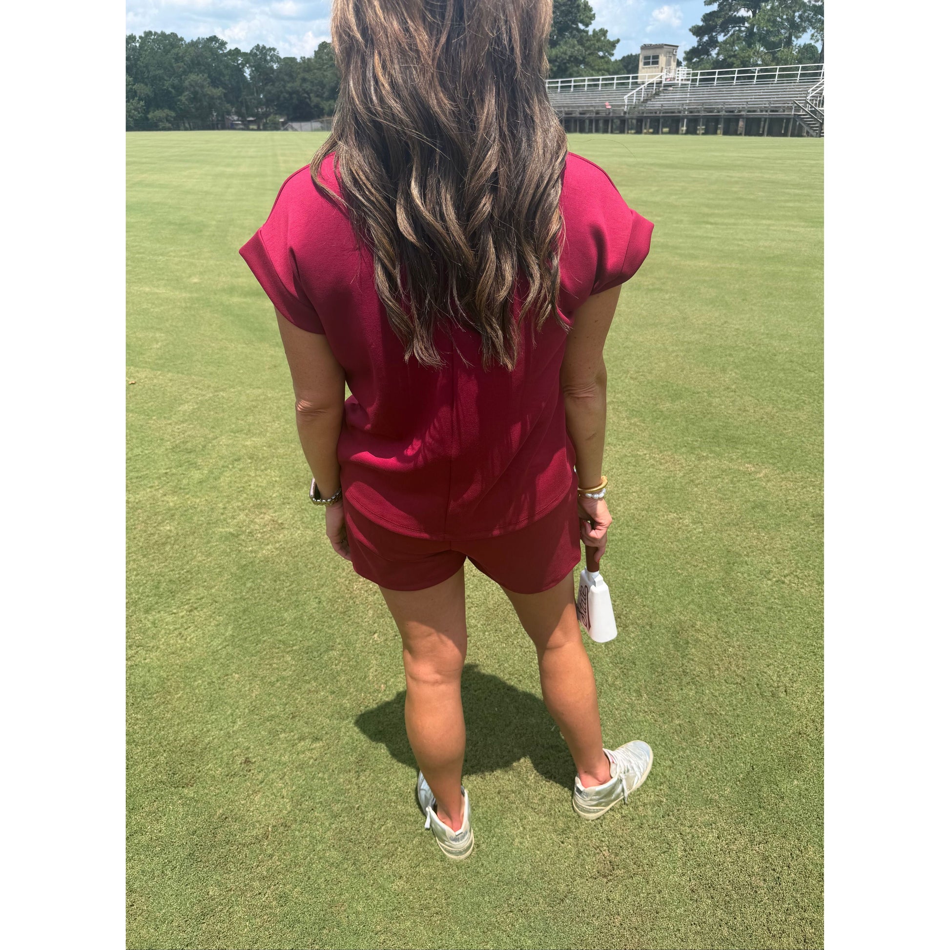 A woman standing on a green grass field wearing a matching magenta short-sleeve top and shorts set, holding a white spray can, with white sneakers.