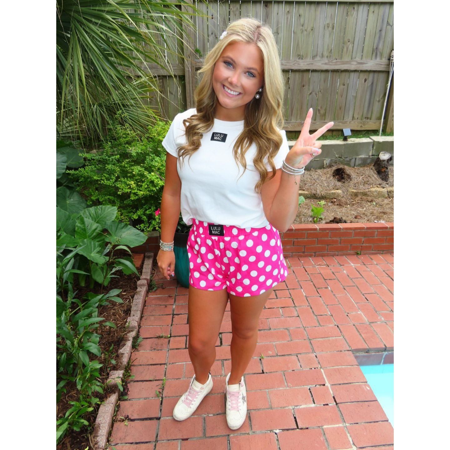 Woman in white t-shirt and pink polka dot shorts standing on a brick patio with plants in the background