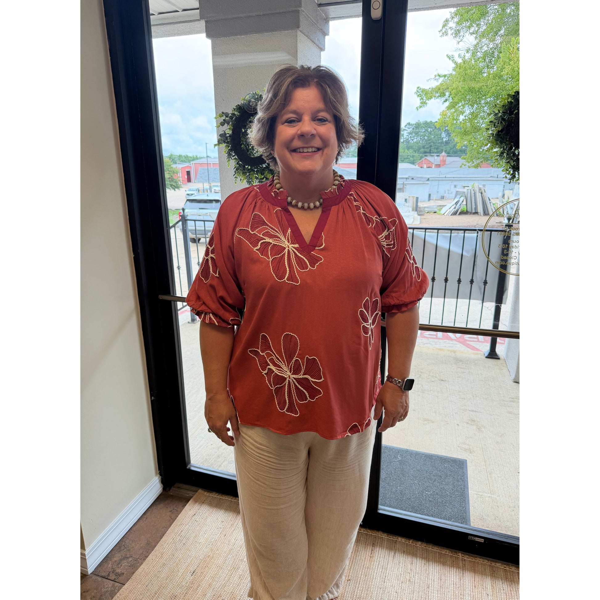 A woman modeling a rust-orange short-sleeve blouse with cream floral embroidery and beige pants, standing in front of a glass door.