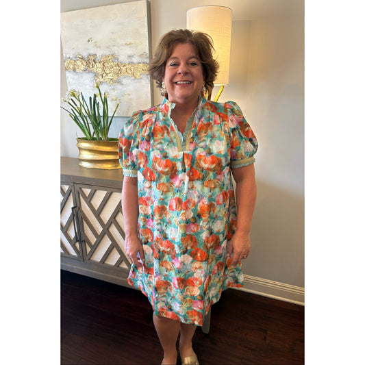 Woman wearing a colorful floral dress standing in a room with a lamp and painting in the background.
