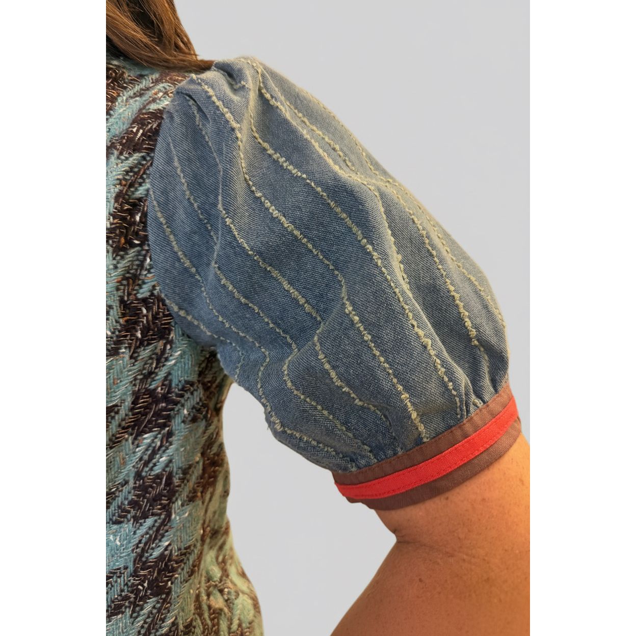 A close-up of a person wearing a blue puff-sleeve top with vertical textured stripes and a red and brown striped cuff, in an indoor setting.