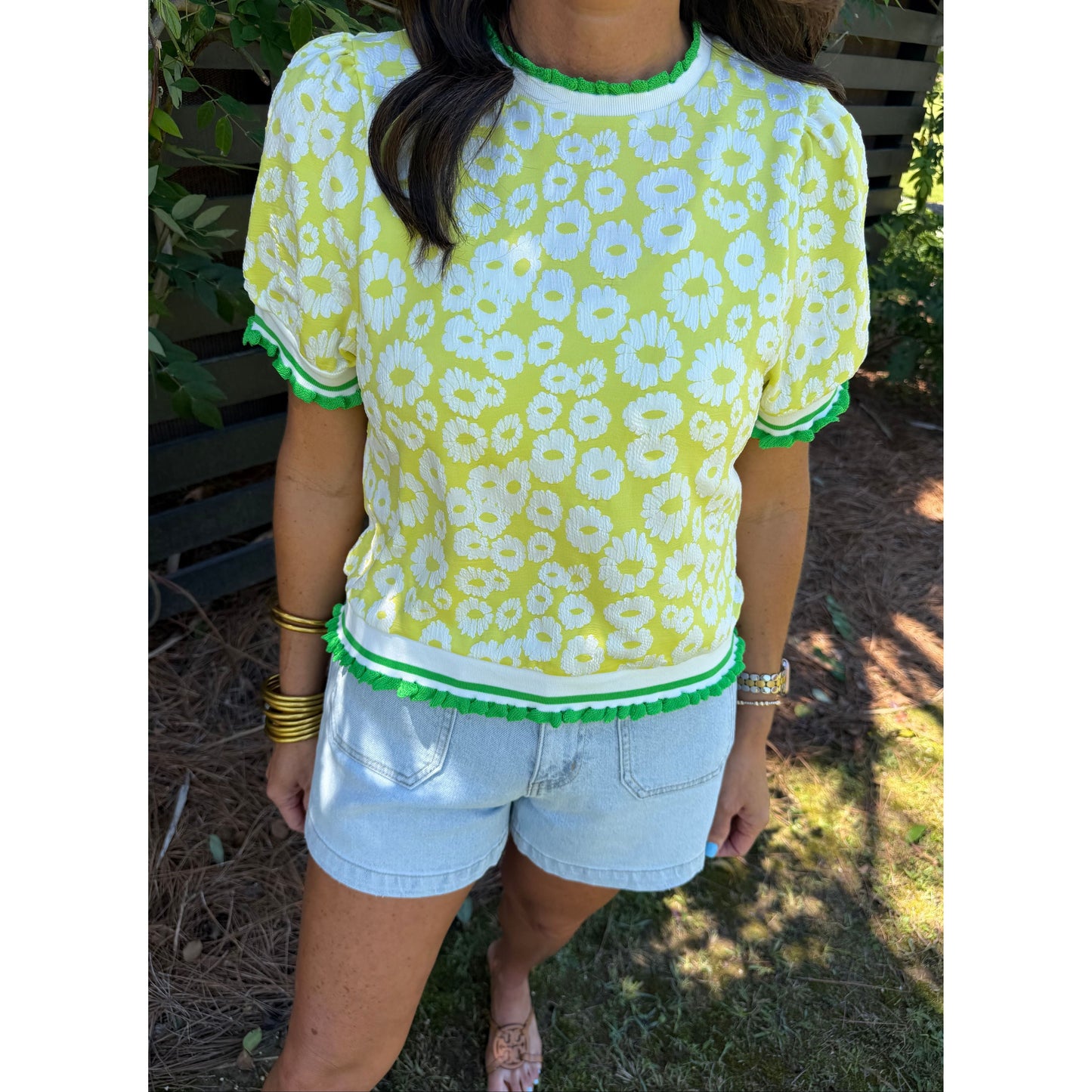 Person wearing a yellow floral blouse with green trim and light blue shorts outdoors.