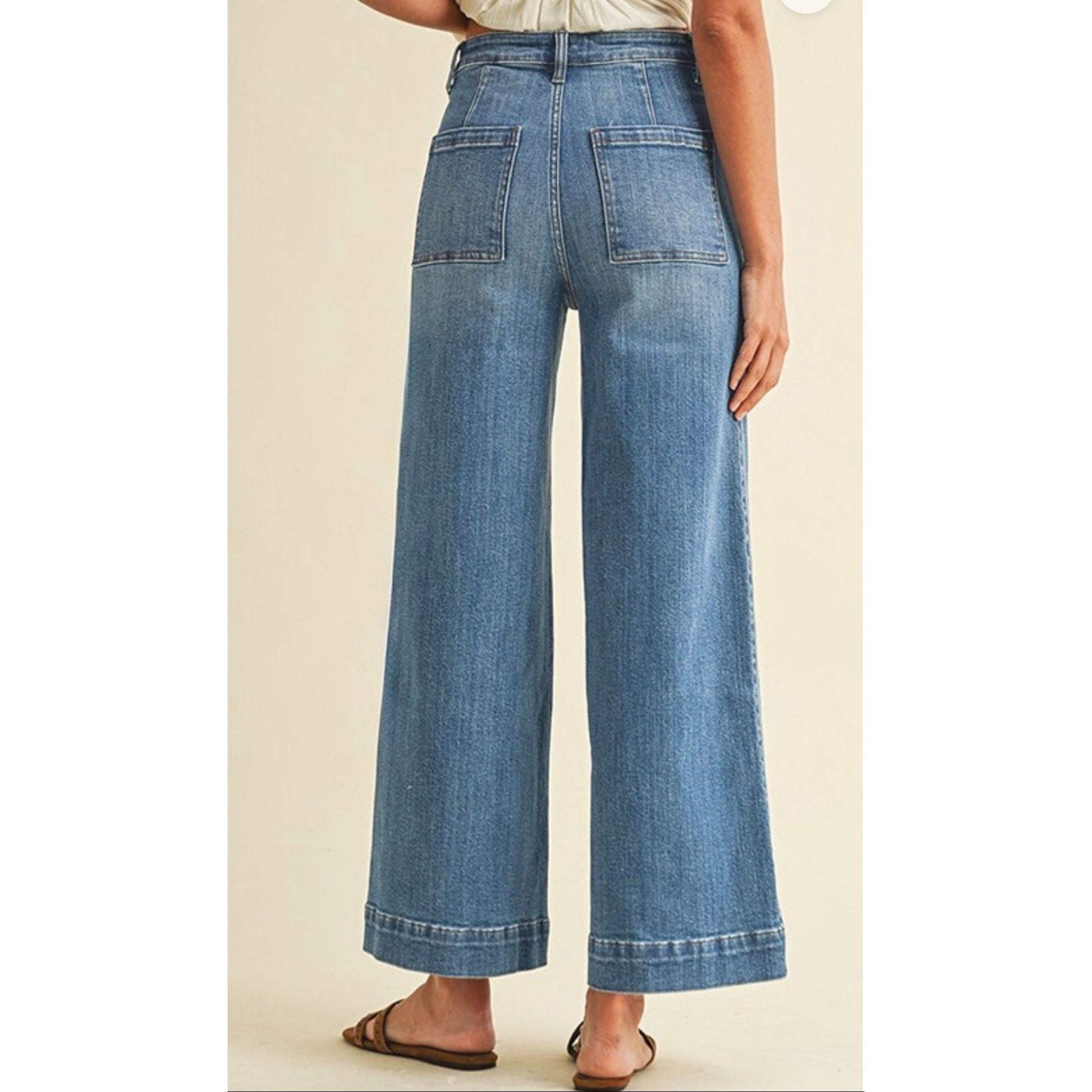 A person shown from the waist down wearing high-waisted, wide-leg blue denim jeans with cuffed hems and brown flat sandals, against a plain beige studio background.