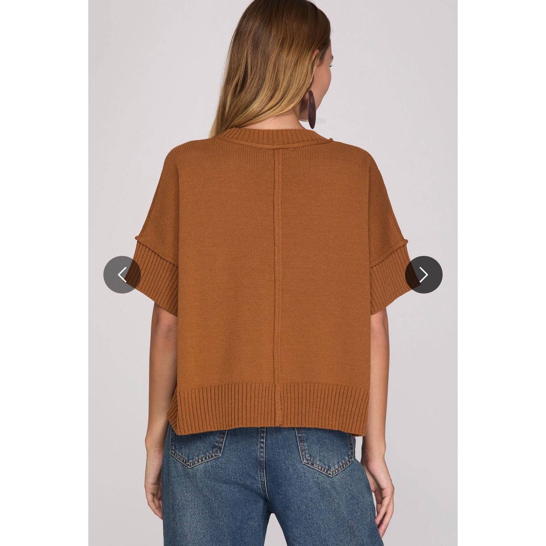 A woman viewed from the back wearing a cropped, short-sleeve knit sweater in a warm camel brown color, paired with blue jeans, against a light gray studio background.