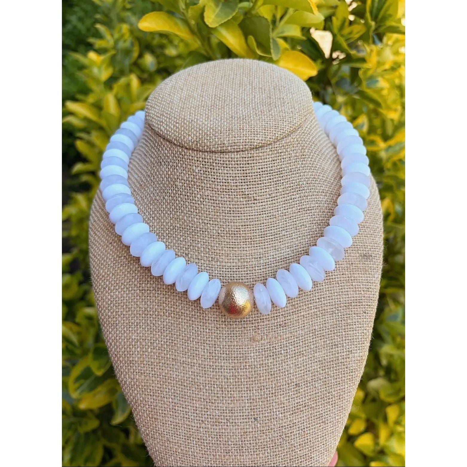 Necklace with white beads and a gold accent on a beige bust against a green leafy background