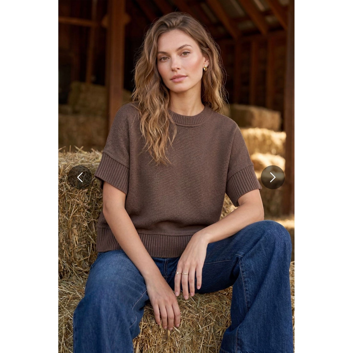 A woman sits on a hay bale wearing a brown short-sleeve knit sweater and blue jeans in a barn setting.